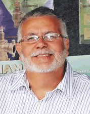 A smiling man with gray hair, a beard, glasses, wearing a striped shirt, in front of a colorful background.