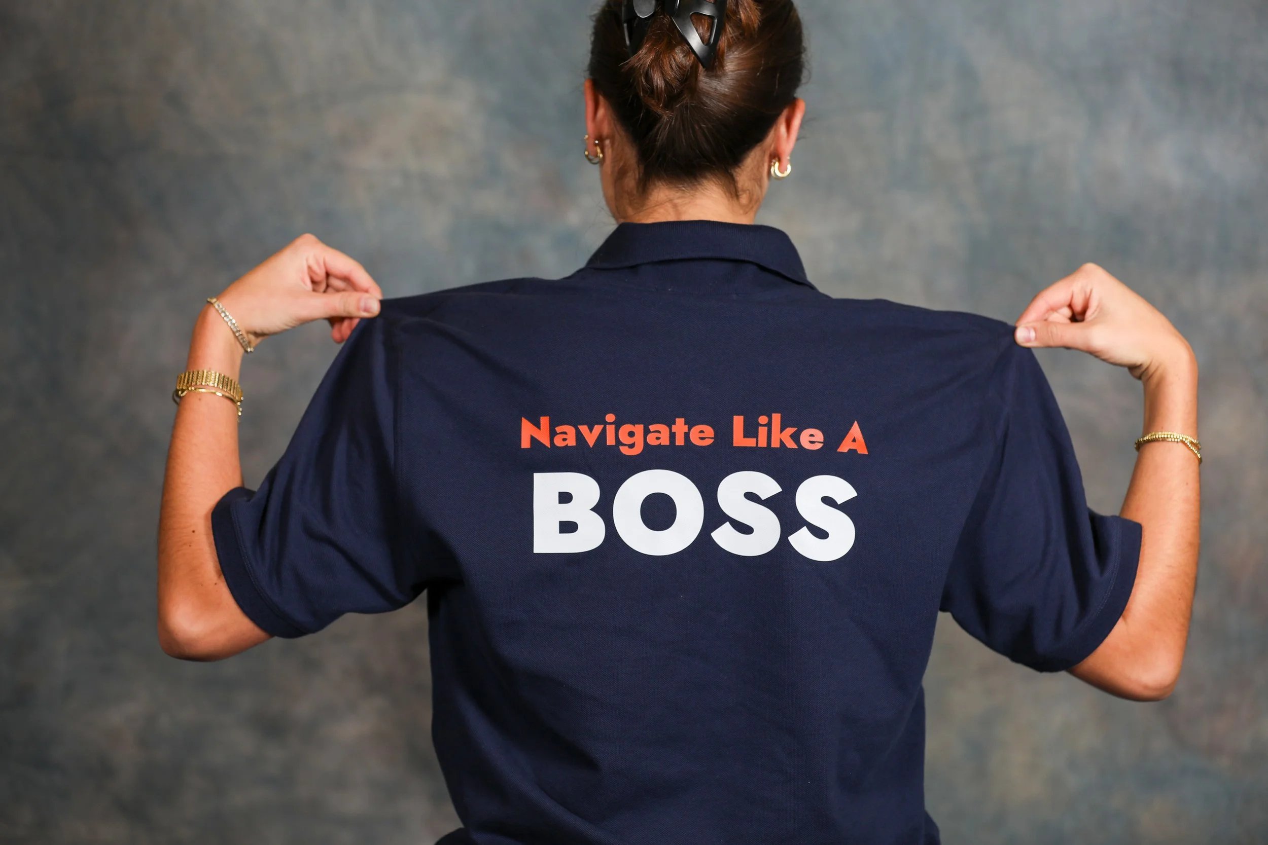 Back of a woman wearing a navy polo shirt with the phrase "Navigate Like A BOSS" printed on it, flexing her arms to show off her muscles, against a textured gray background.