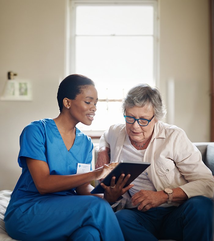 A nurse in blue scrubs sitting with an elderly woman, showing her something on a tablet in a well-lit room.