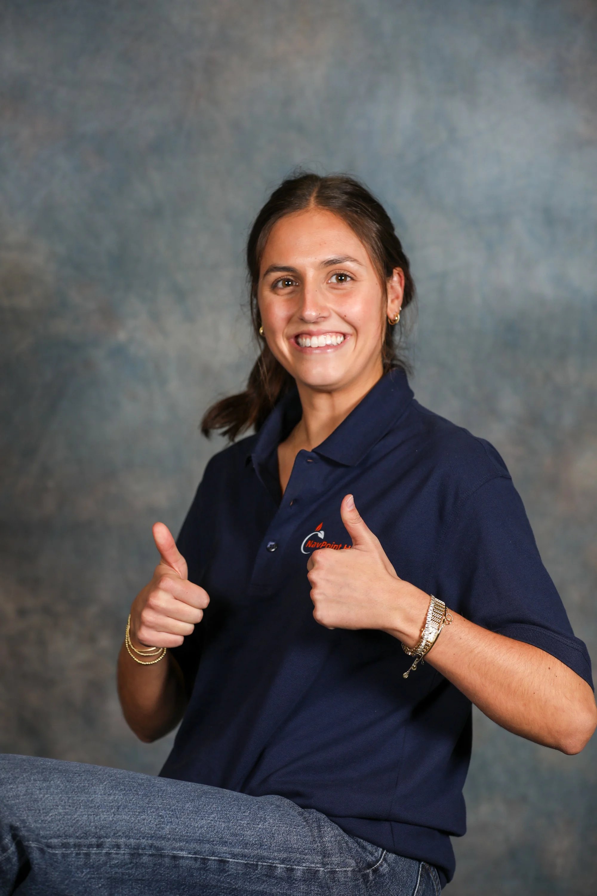 A young woman smiling and giving two thumbs up, wearing a navy blue polo shirt with a company logo, sitting against a gray backdrop.