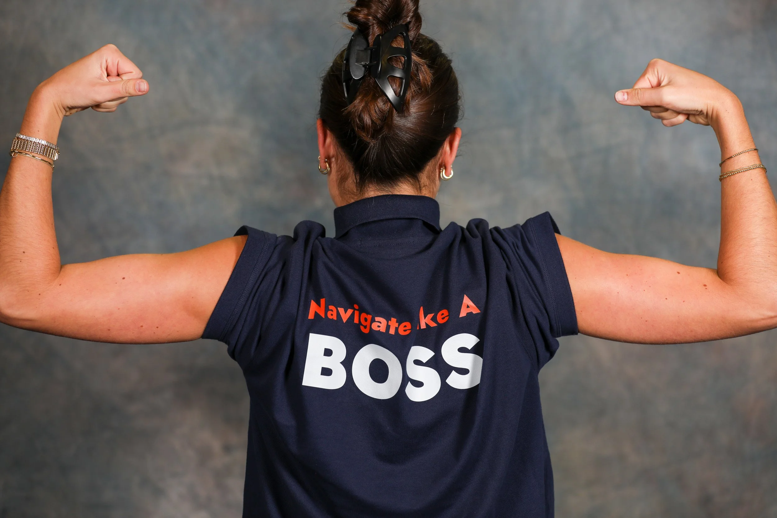 Back view of a woman wearing a navy sports jersey with the words 'Navigate Like A BOSS' printed on it, flexing her biceps.