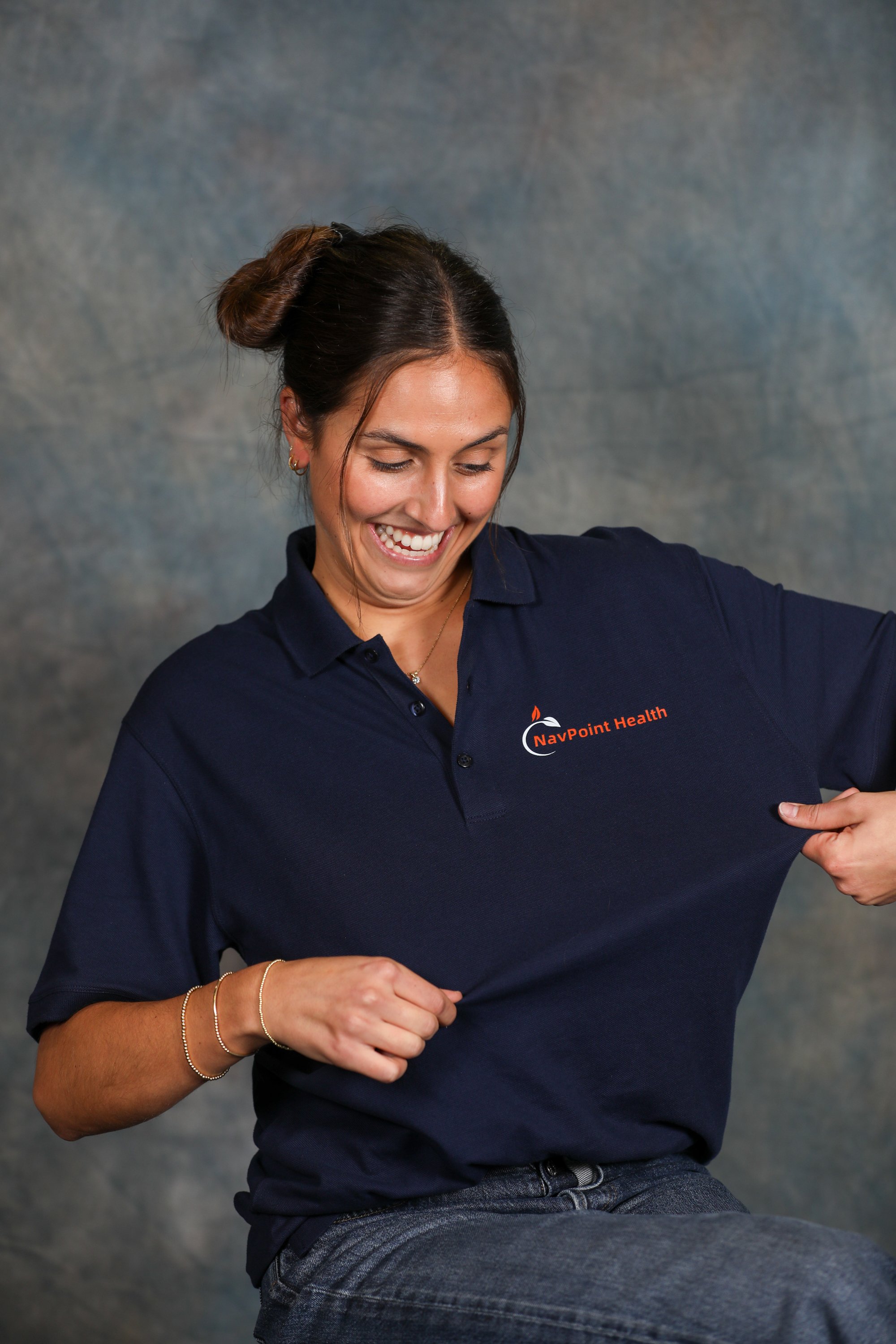 A woman smiling and pulling her navy blue polo shirt to reveal the orange NavPoint Health logo on the chest.