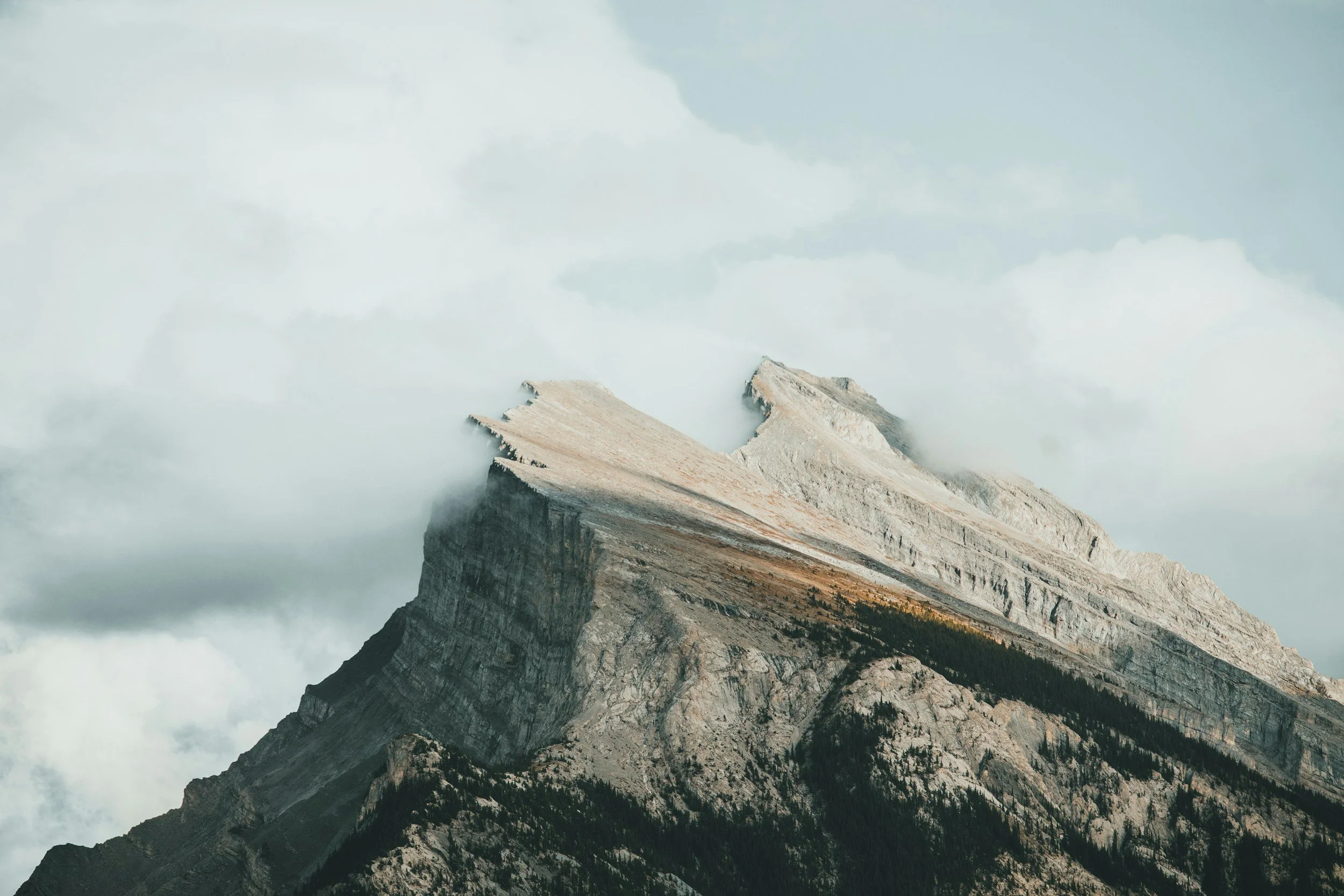 A mountain peak with a flat top, partly shrouded in clouds, surrounded by smaller mountains and forested slopes.