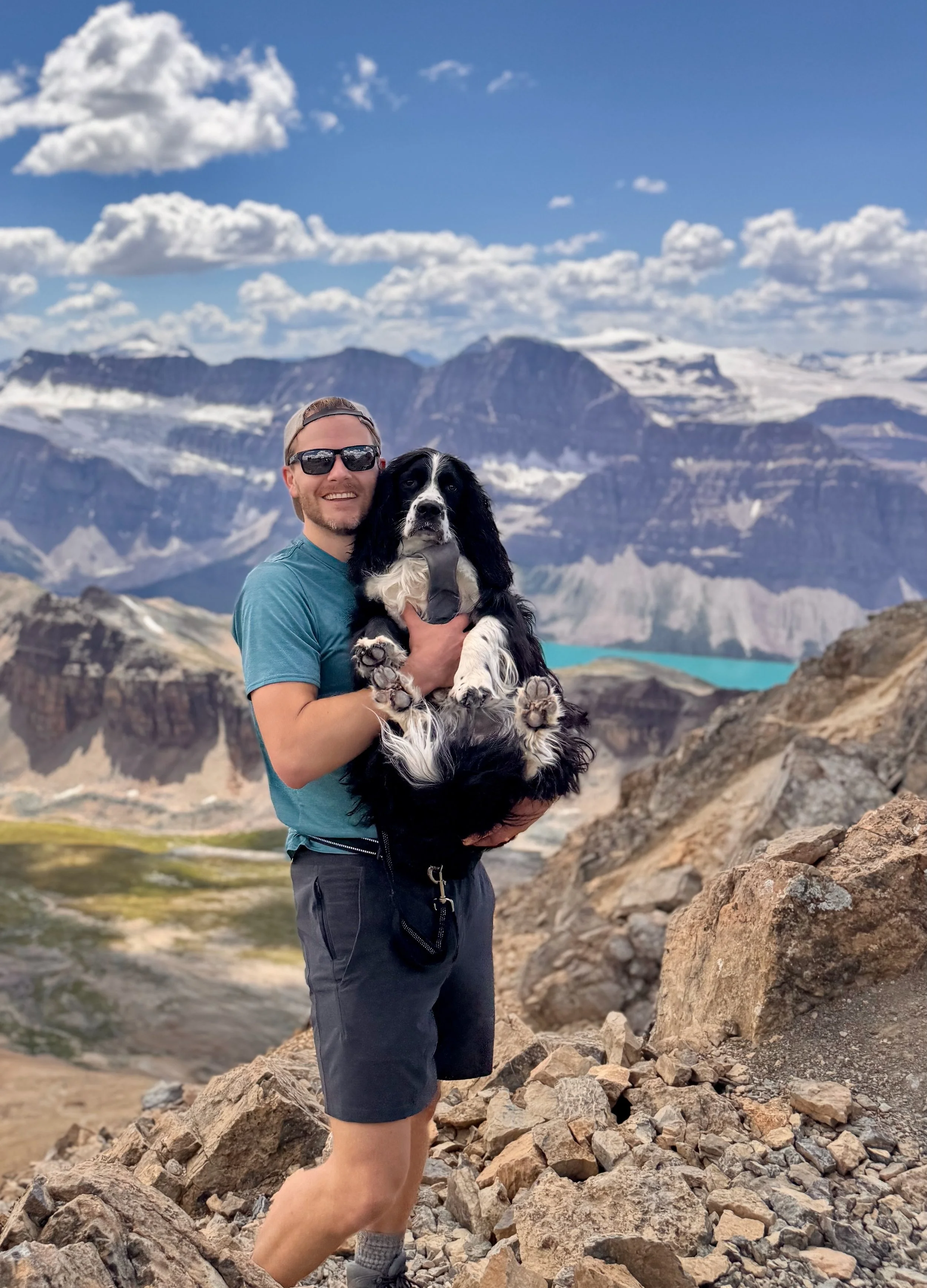 Man wearing sunglasses and a blue t-shirt holding a black and white dog in a mountainous landscape with snow-capped peaks and a lake in the background.