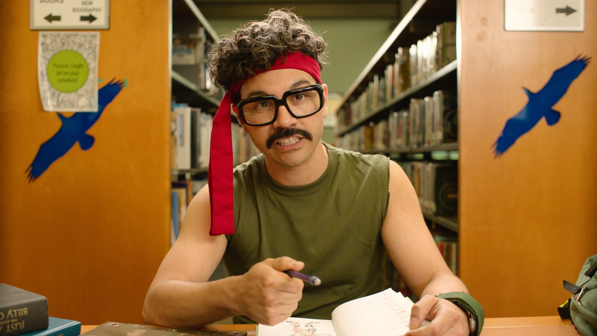 A man with curly hair, glasses, and a red headband sitting at a table in a library, holding a pen and looking at the camera.