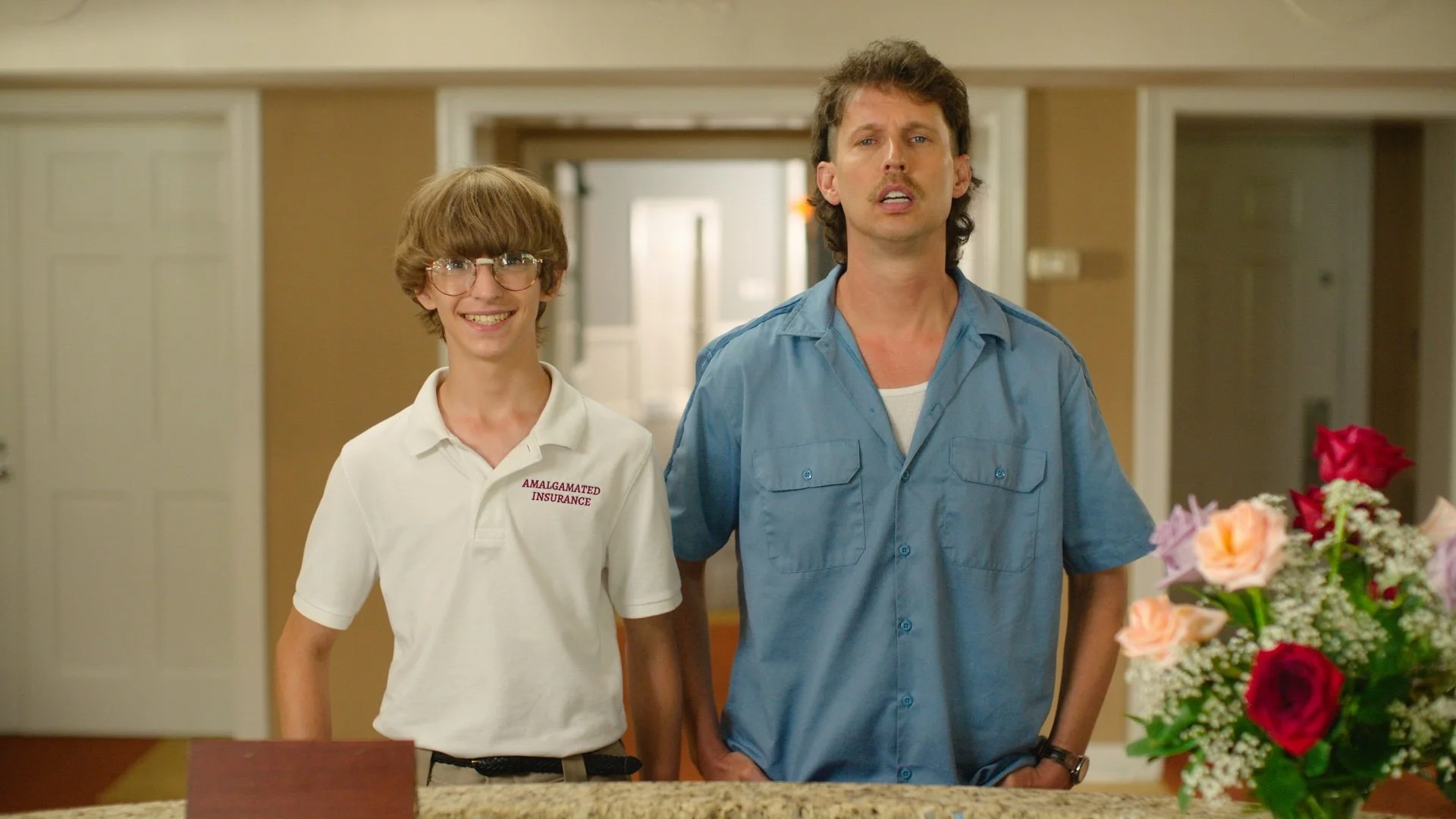 Two men, one younger with glasses and a white polo shirt that says 'Amalgamated Insurance', and one older with a blue shirt, are walking into a room with a floral arrangement in the foreground, inside a house.