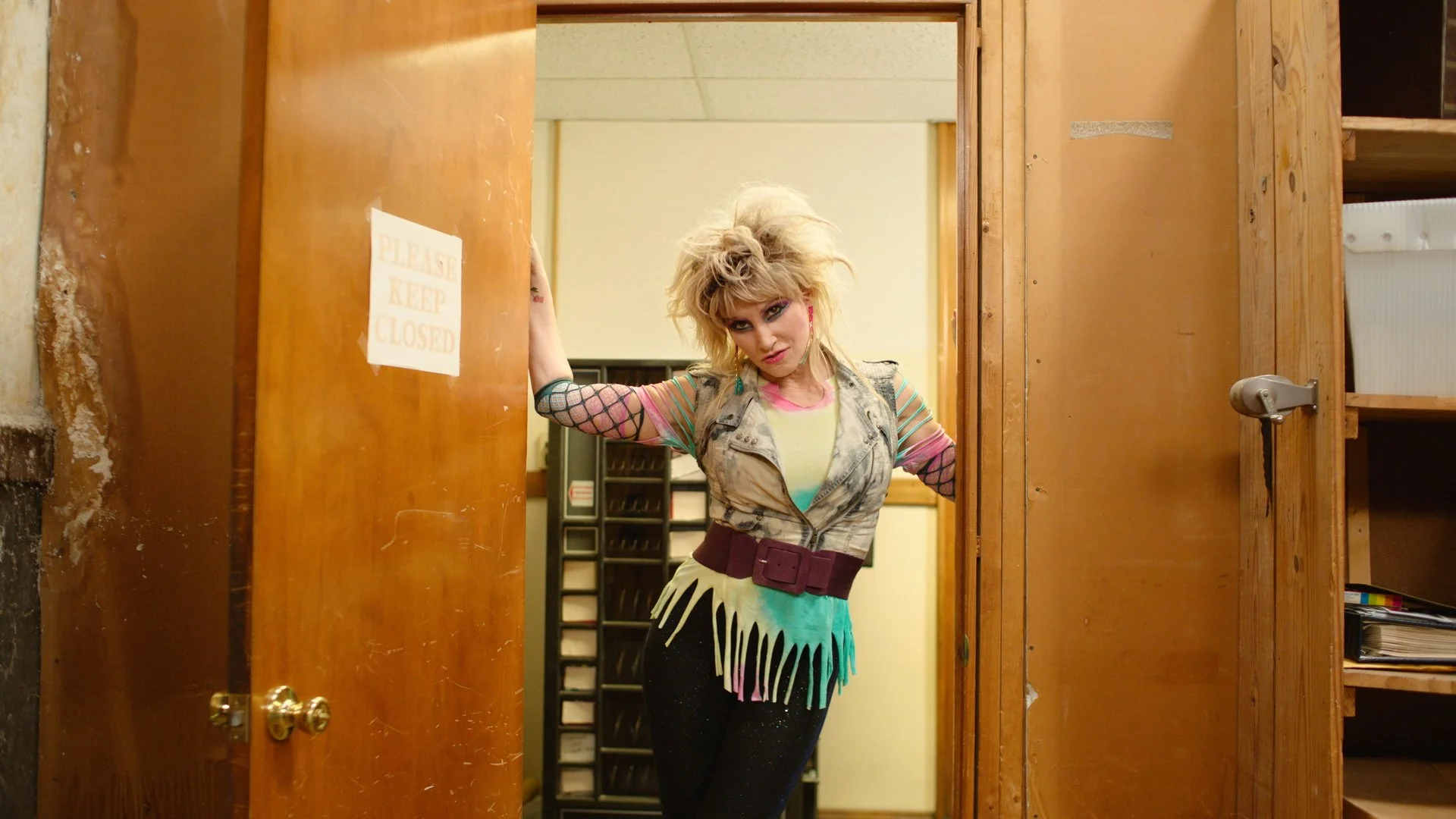 A woman with wild blonde hair and colorful makeup standing in a doorway, wearing a eclectic outfit with fishnet sleeves, a vest, a colorful dress, and a large belt, in an indoor setting with wooden walls.