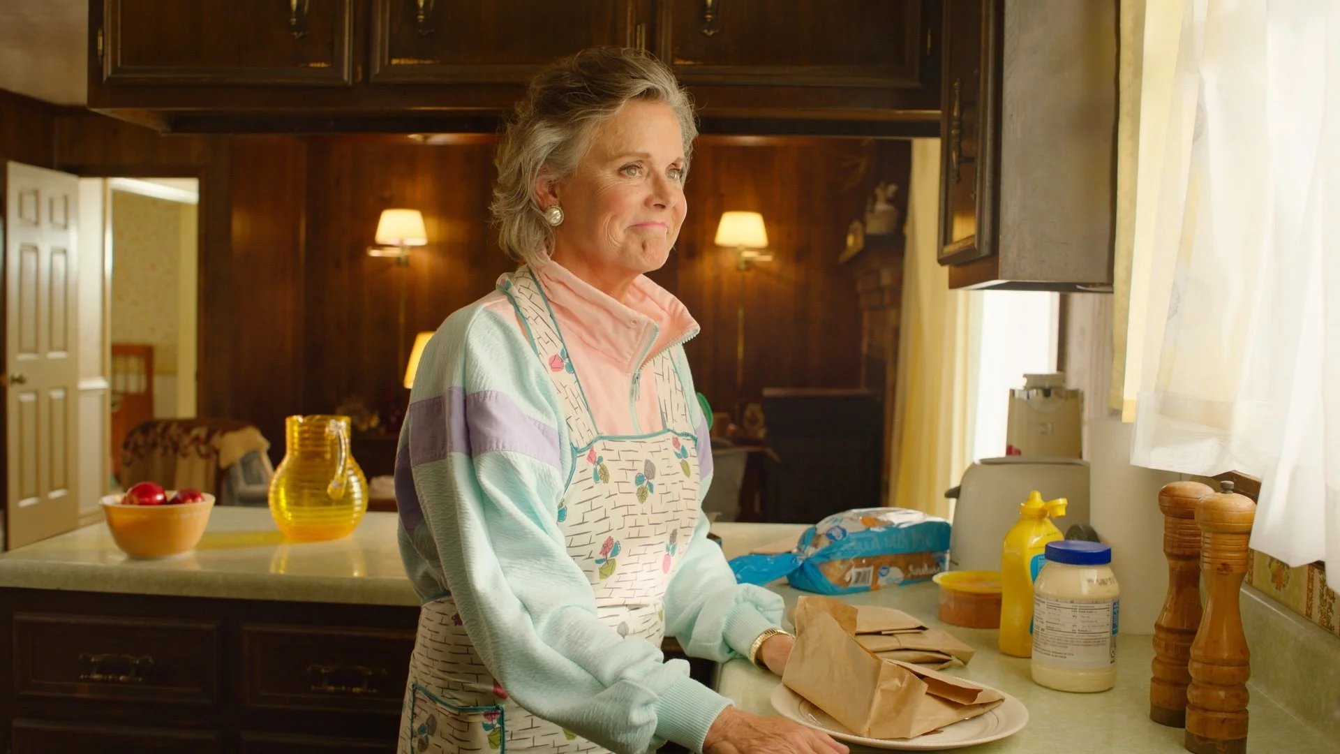 An elderly woman with gray hair, wearing pearl earrings, a pastel striped sweater, and a floral apron, stands in a kitchen near a counter with paper bags, condiments, and a container of sour cream, looking thoughtfully to her right. The kitchen has d
