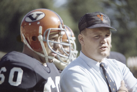 A football coach in a white shirt and tie standing next to a player in a football uniform and helmet, with other players in the background.