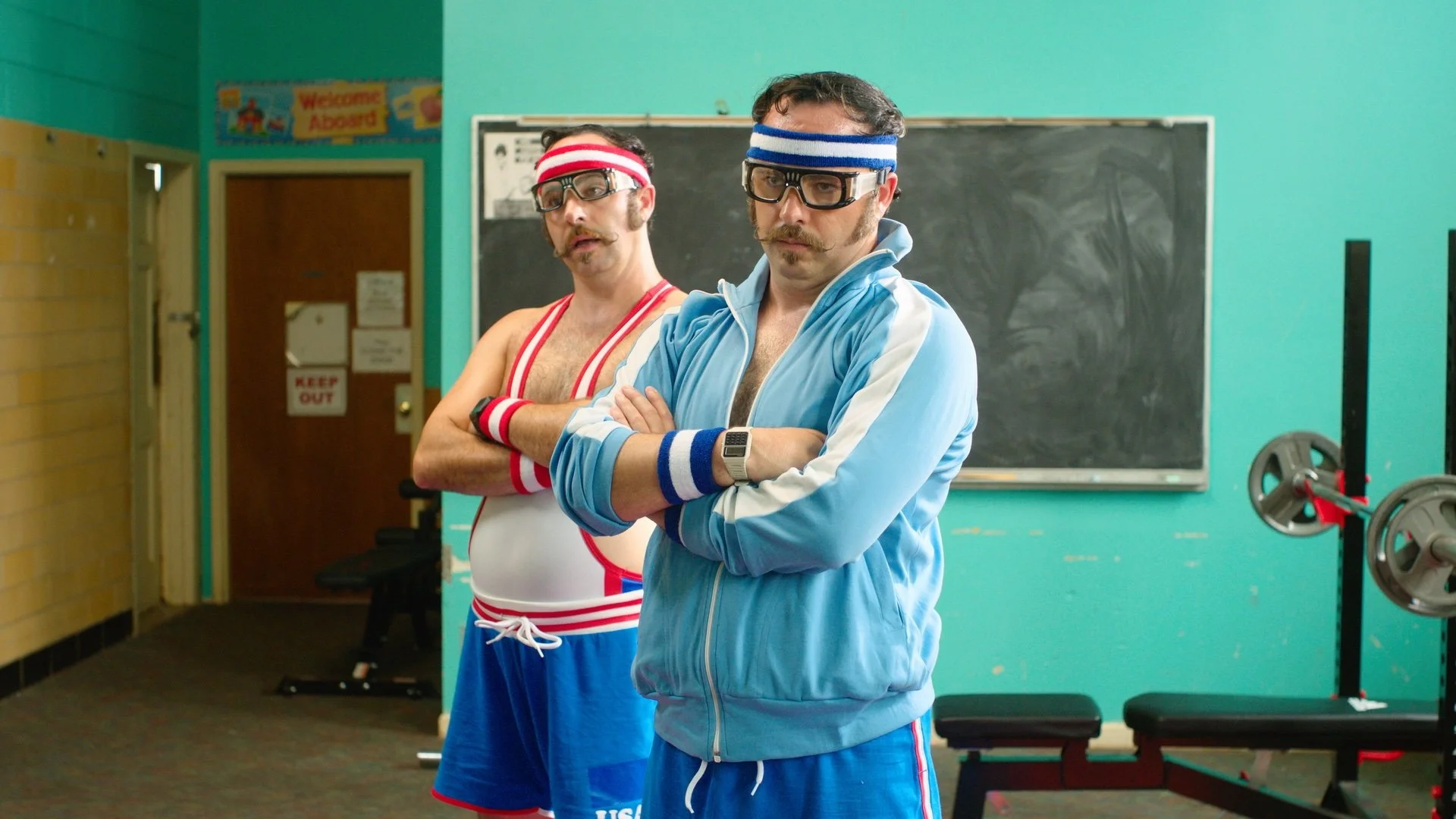 Two men dressed in vintage-style sportswear, with headbands and goggles, standing in a gym locker room.