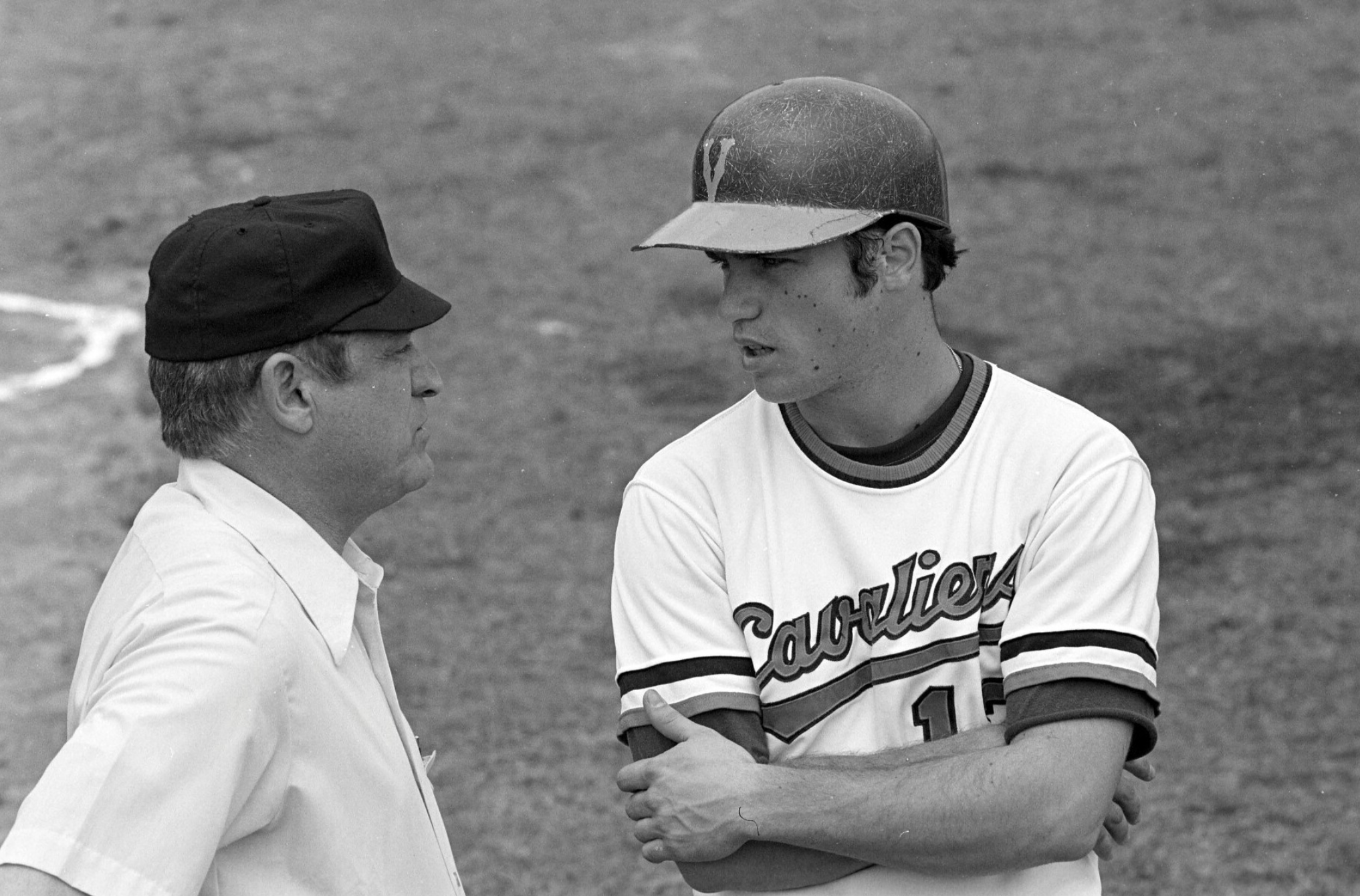 Two men are having a conversation on a baseball field, one wearing a baseball uniform and a helmet, the other in a white shirt and cap.