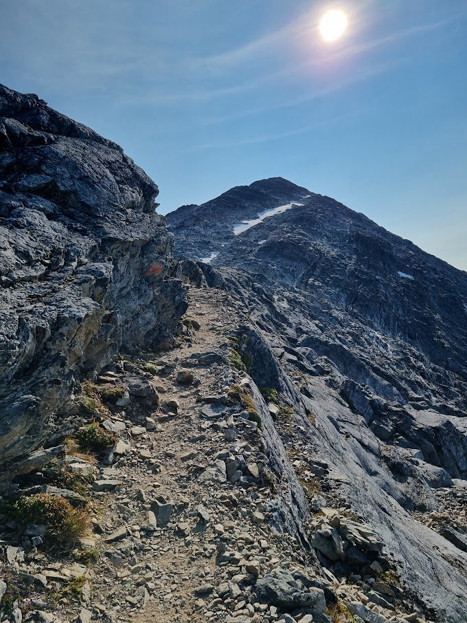 A rocky mountain trail with steep, rugged terrain leading up to a mountain peak under a bright sun and clear blue sky.