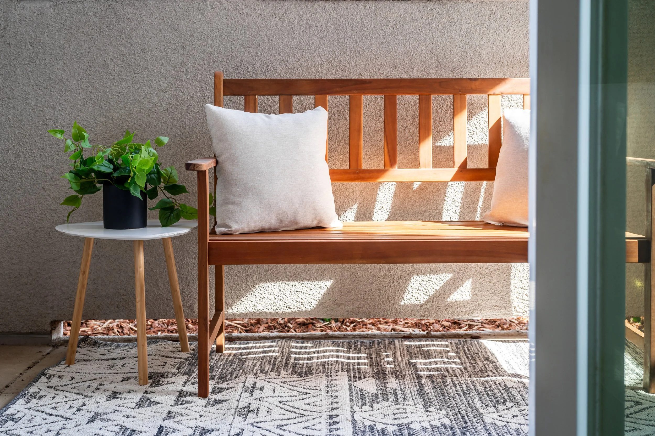 A porch with a wooden bench with two white pillows, a small side table with a potted green plant, a patterned outdoor rug, a beige textured wall, and sunlight casting shadows.