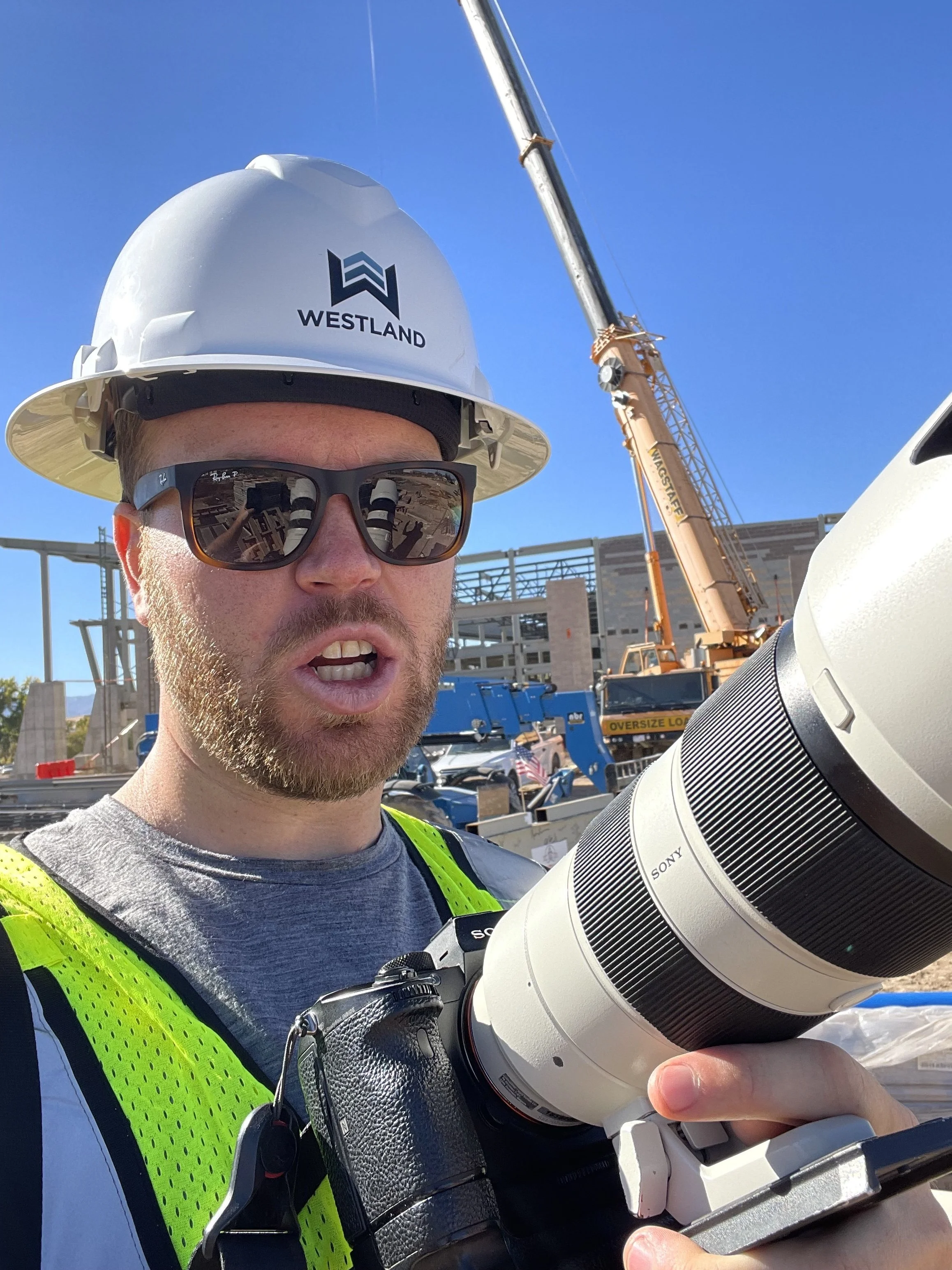 Berin Klawiter in a Westland hard hat and safety vest holding a Sony telephoto lens at an active construction site — architectural photography on location