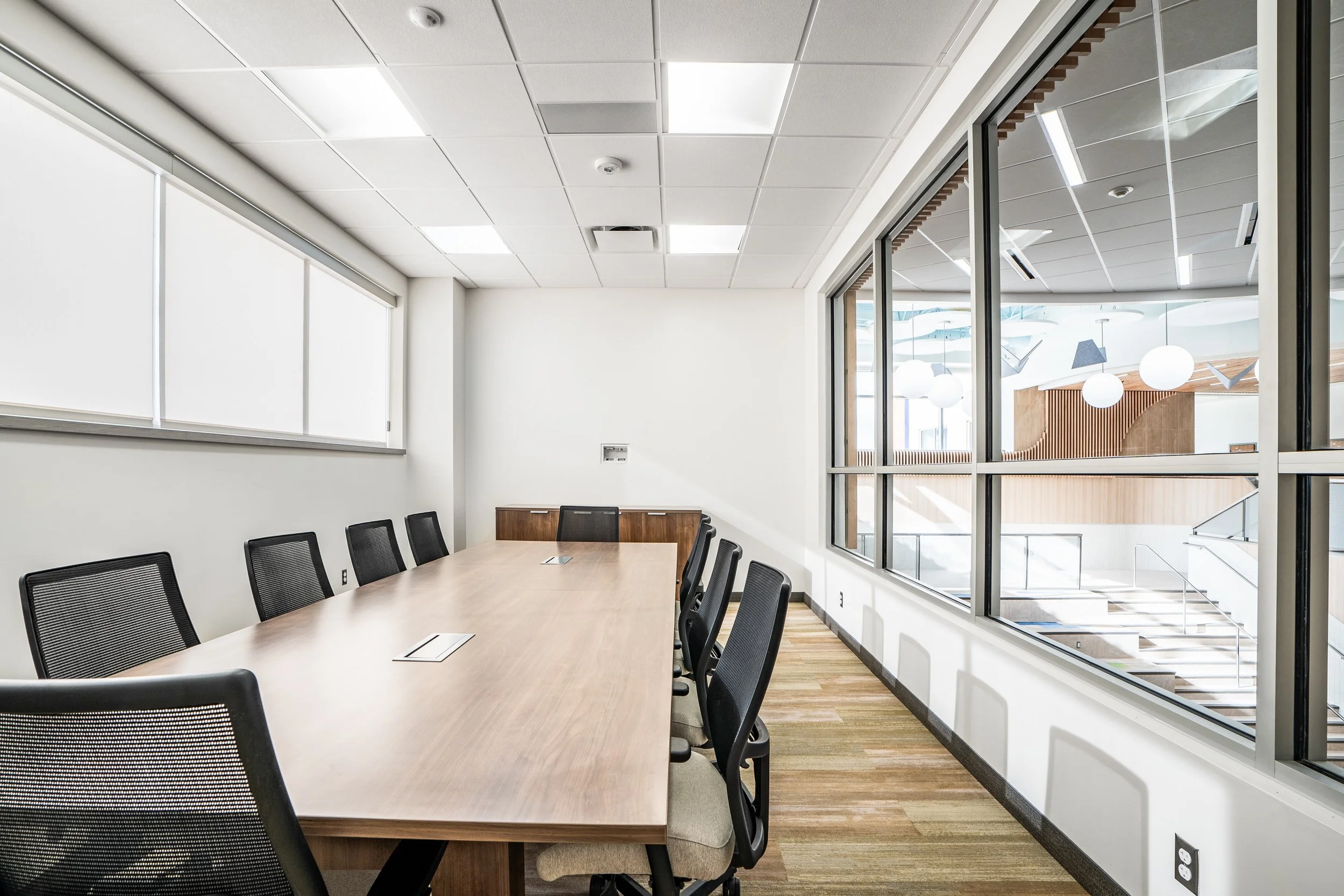 Modern office conference room with a long wood table, mesh chairs, and an interior window overlooking a staircase