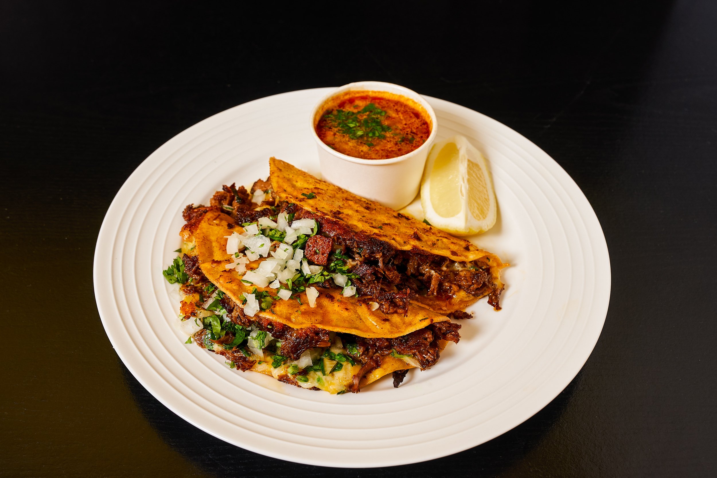 Quesabirria tacos with onion and cilantro served with a cup of consomé and lime wedges on white plate — restaurant menu photography by BK Captured