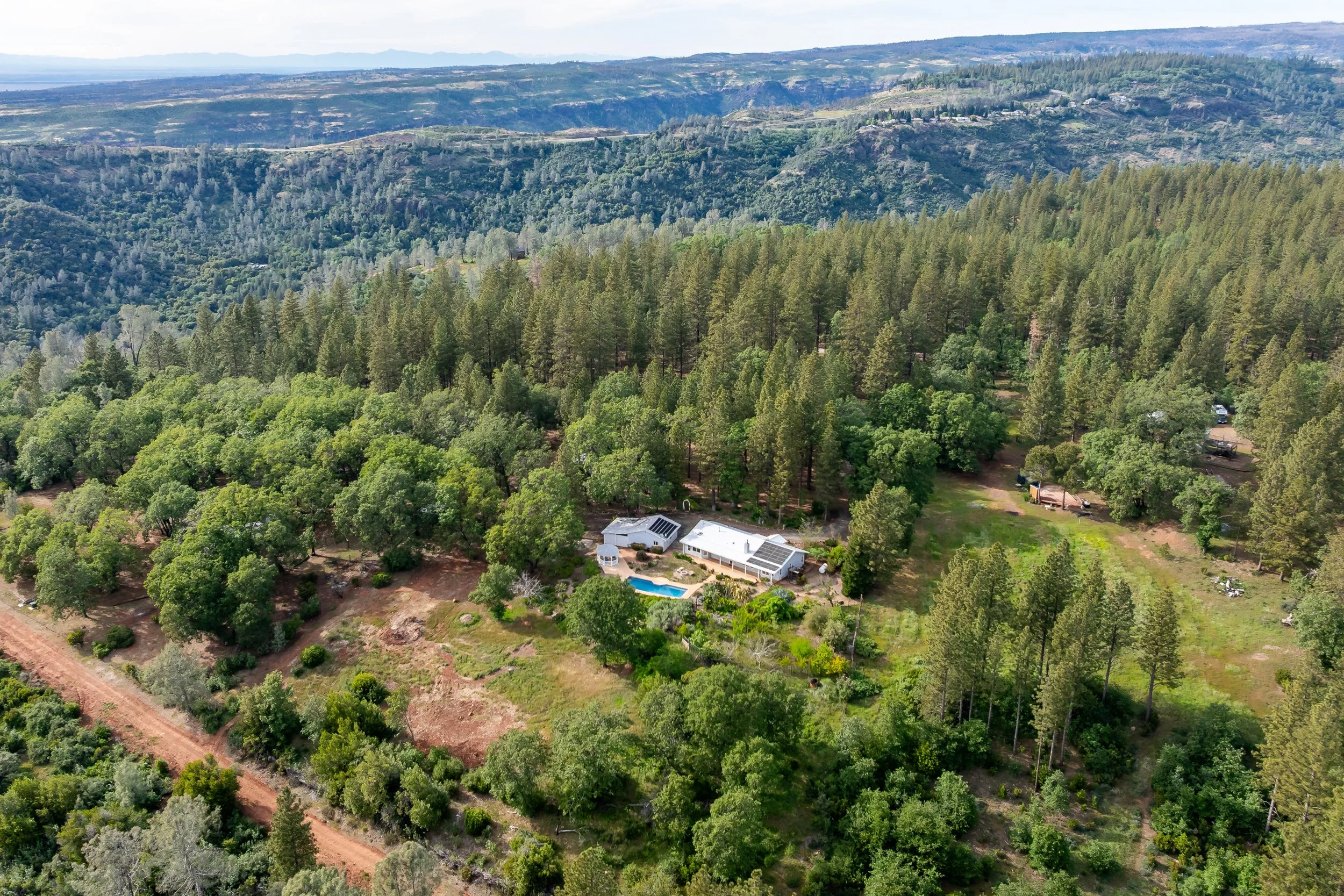 Aerial view of green forested hills with a white house, swimming pool, and solar panels in the yard. Distant mountains and a cloudy sky in the background.