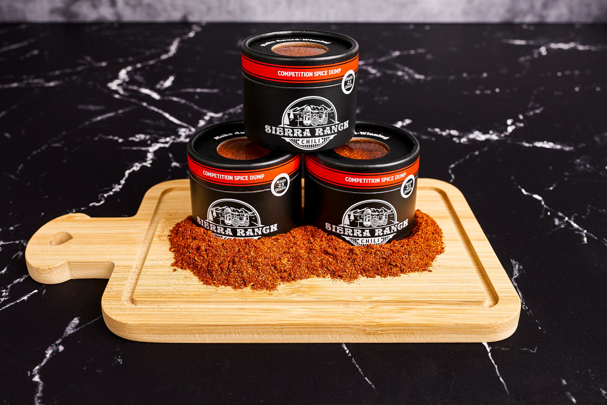 Three Sierra Ranch Competition Spice Dump canisters stacked on a cutting board with spice pile on black marble — food product photography by BK Captured