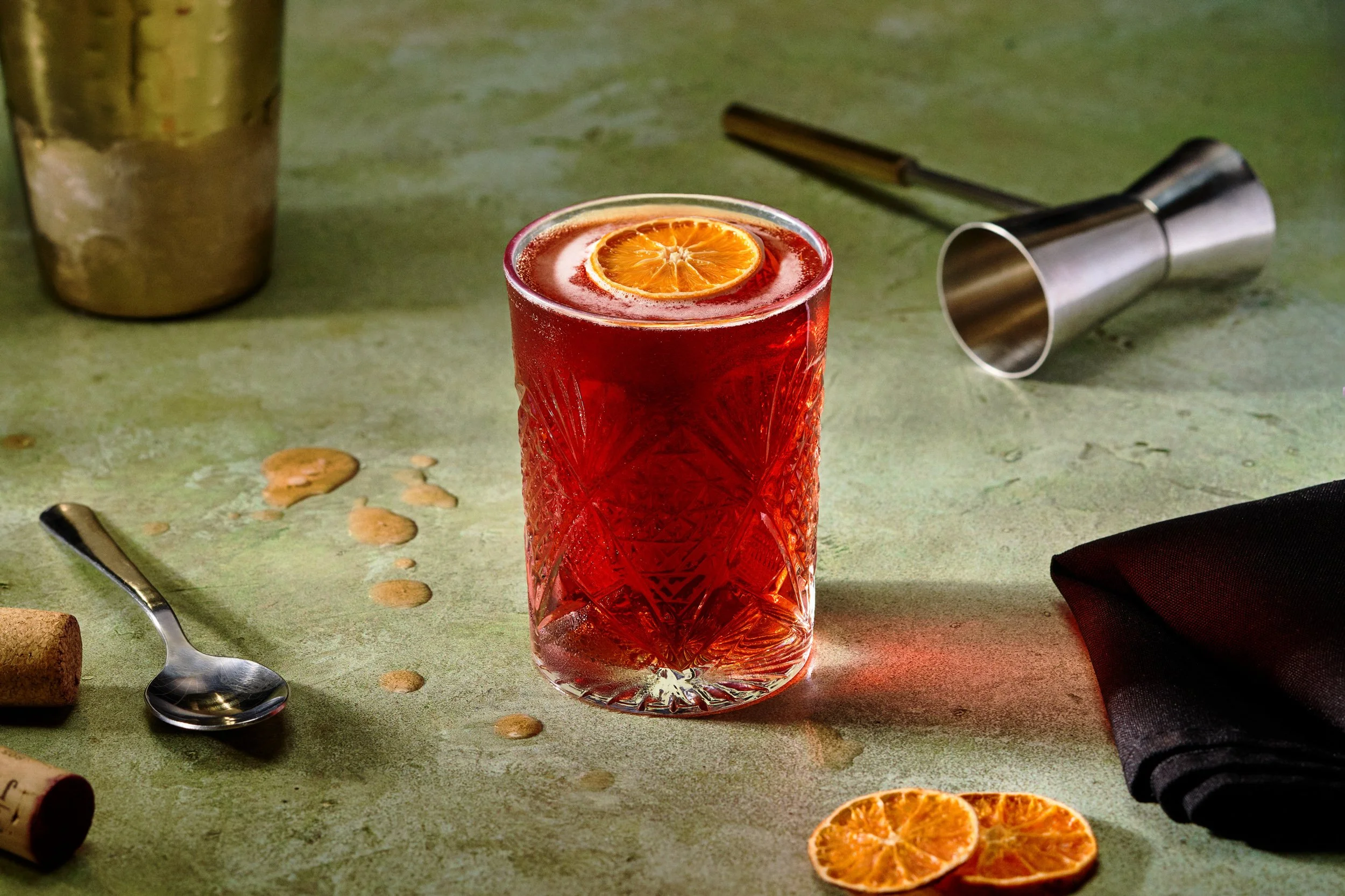 A glass of bright red beverage garnished with a slice of orange, placed on a green surface with scattered orange slices, a metal cocktail shaker lying on its side, a spoon, wine cork, and a folded black napkin nearby.