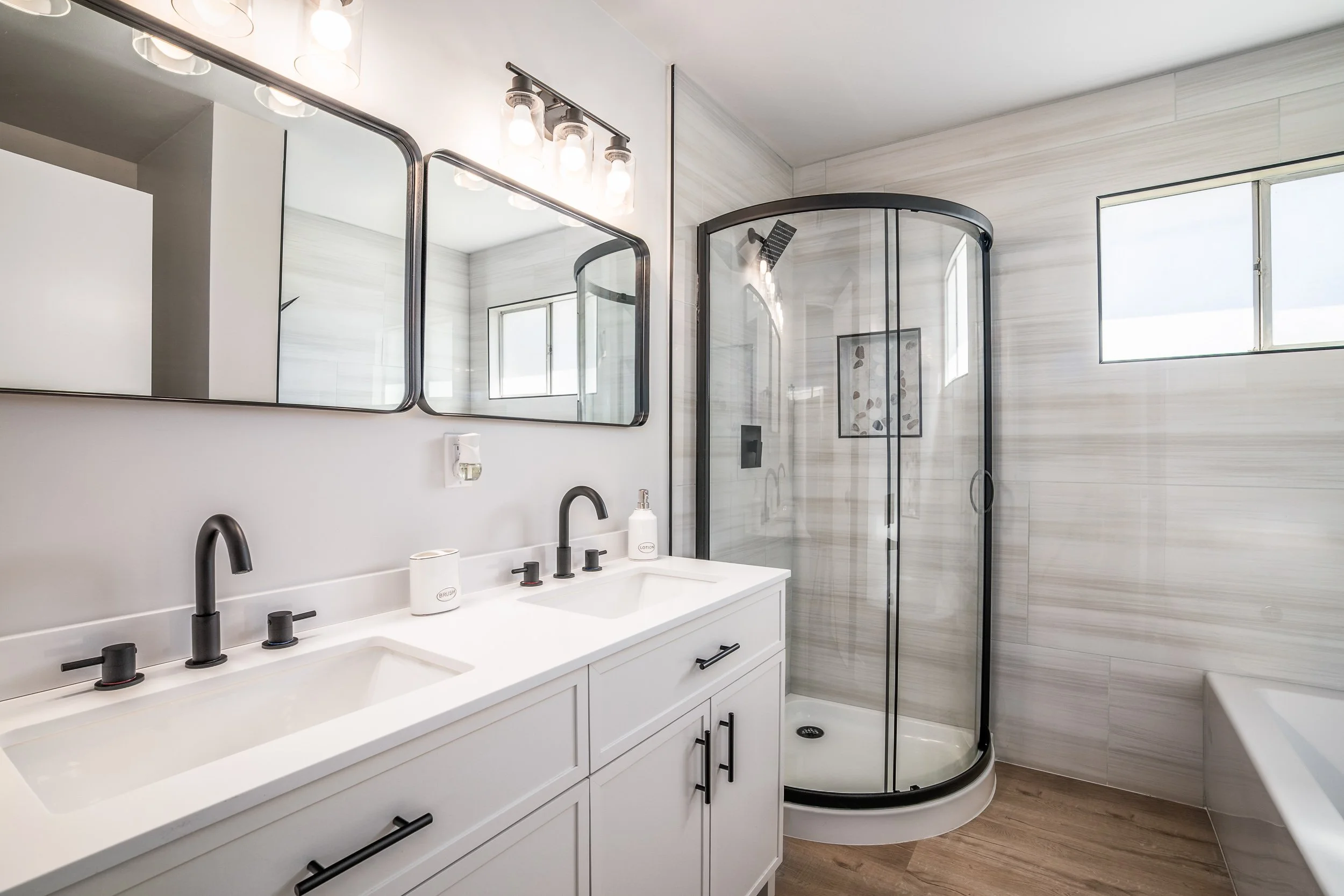 Renovated bathroom with a dual white vanity, black fixtures, and a curved glass corner shower