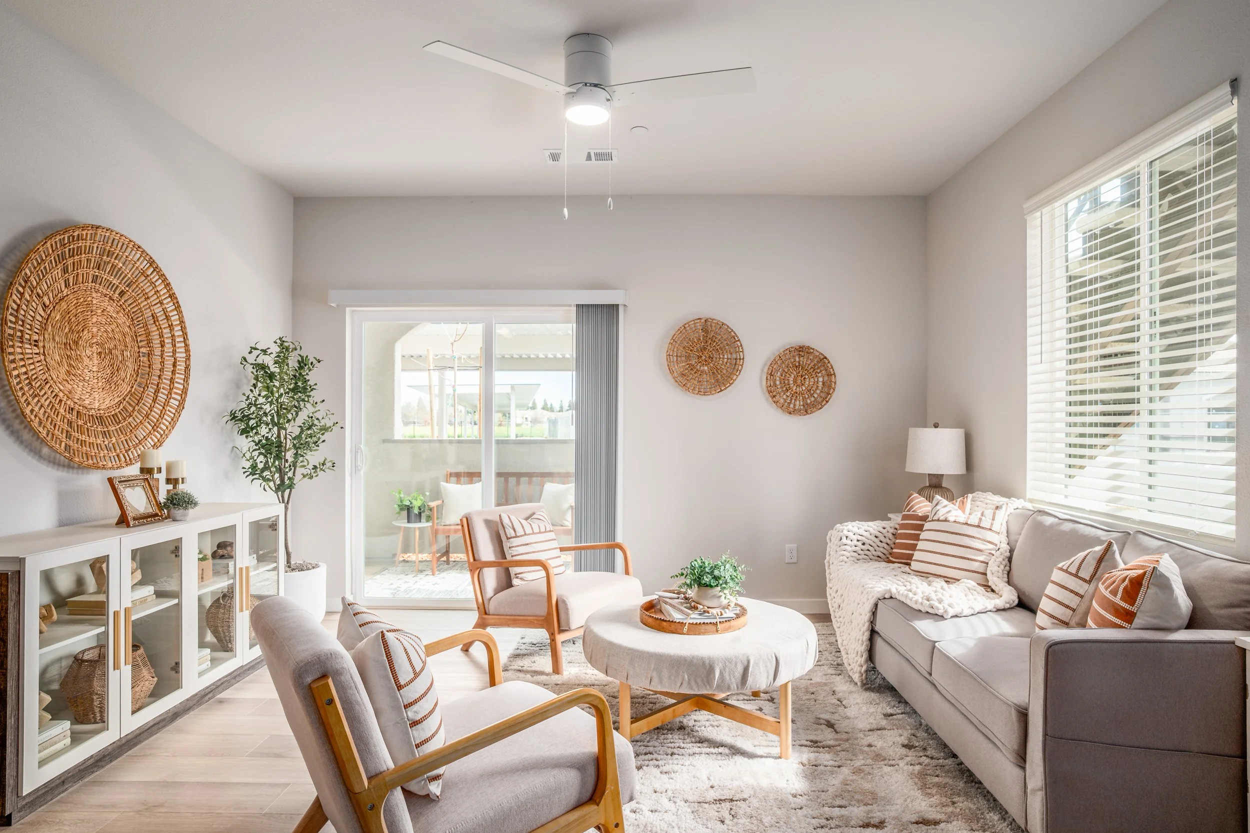 A bright living room with white walls, a ceiling fan, and large windows with blinds. The room features a white sideboard with decor, a green potted plant, a beige armchair, a white coffee table with a plant, and a light gray sofa with striped pillows