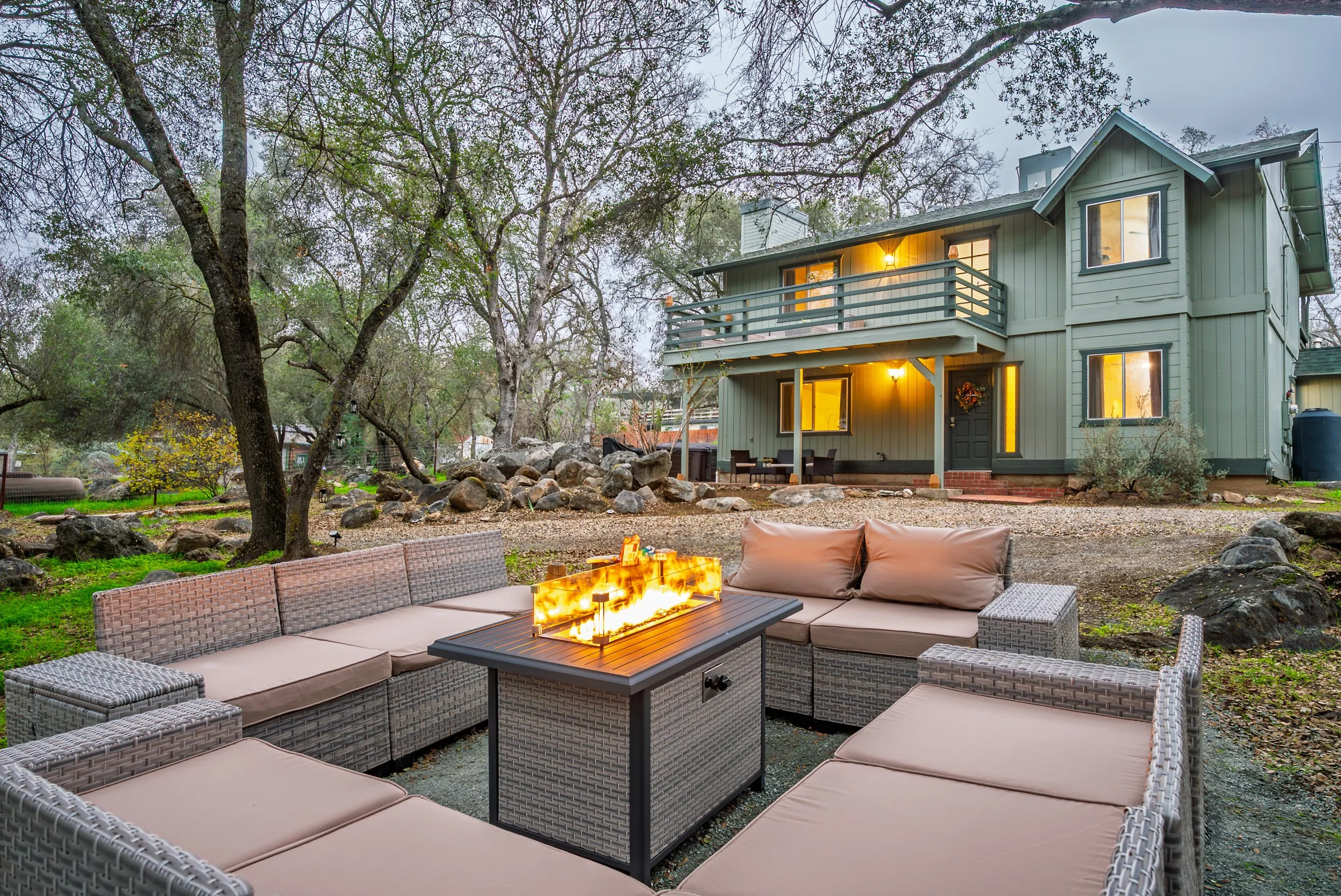 A backyard scene with a firepit, outdoor seating, trees, and a two-story house with a balcony, illuminated windows, and outdoor lighting at dusk.
