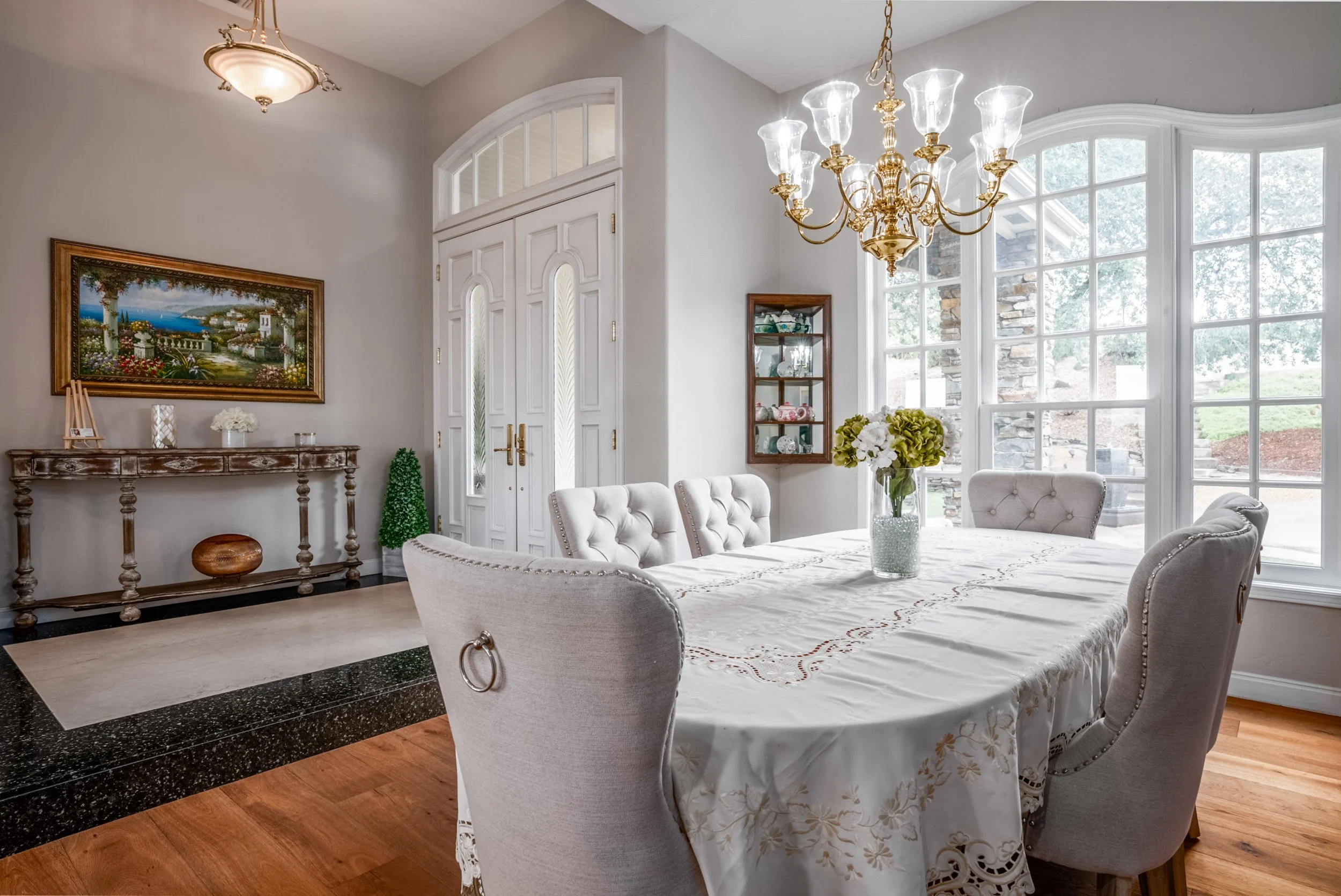 Formal dining room with brass chandelier, tufted linen chairs, and tall arched windows