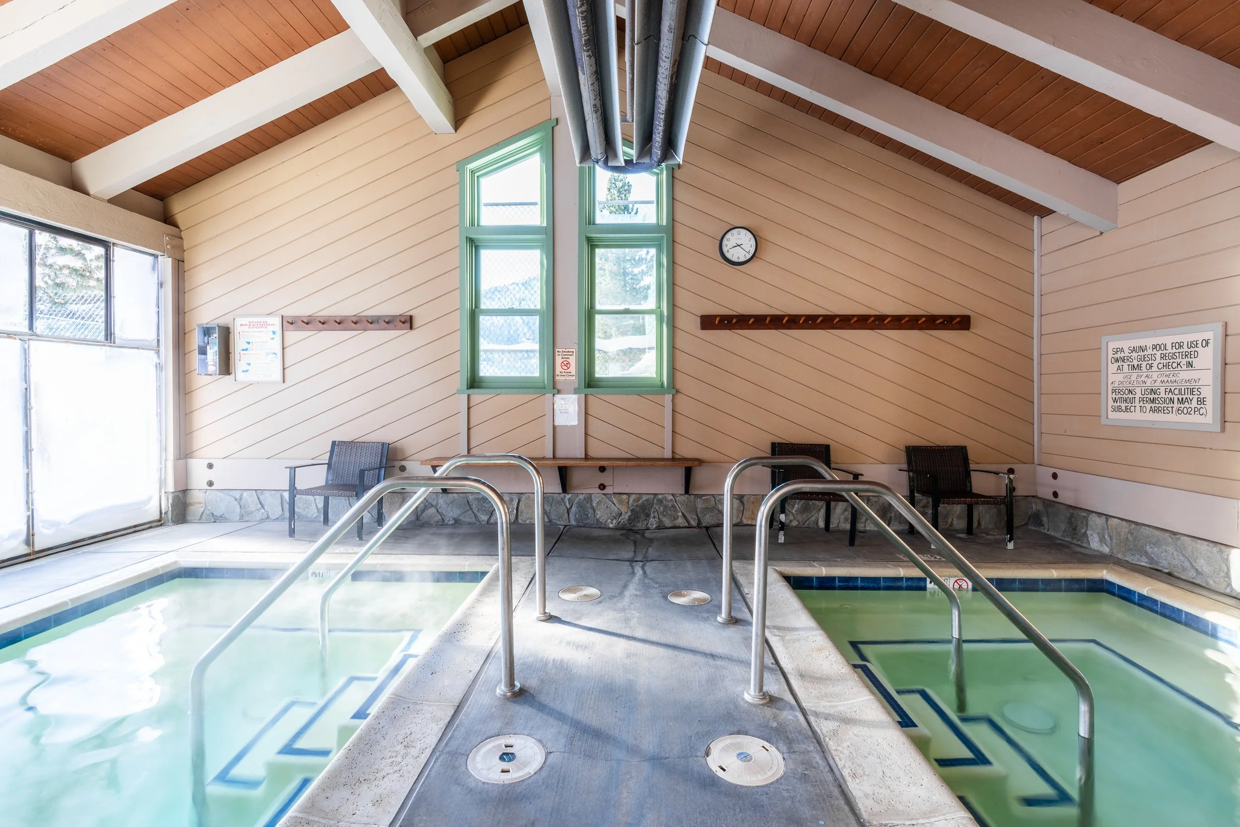 Indoor pool area with two hot tubs, green trim windows, beige wooden wall panels, clocks, and chairs.