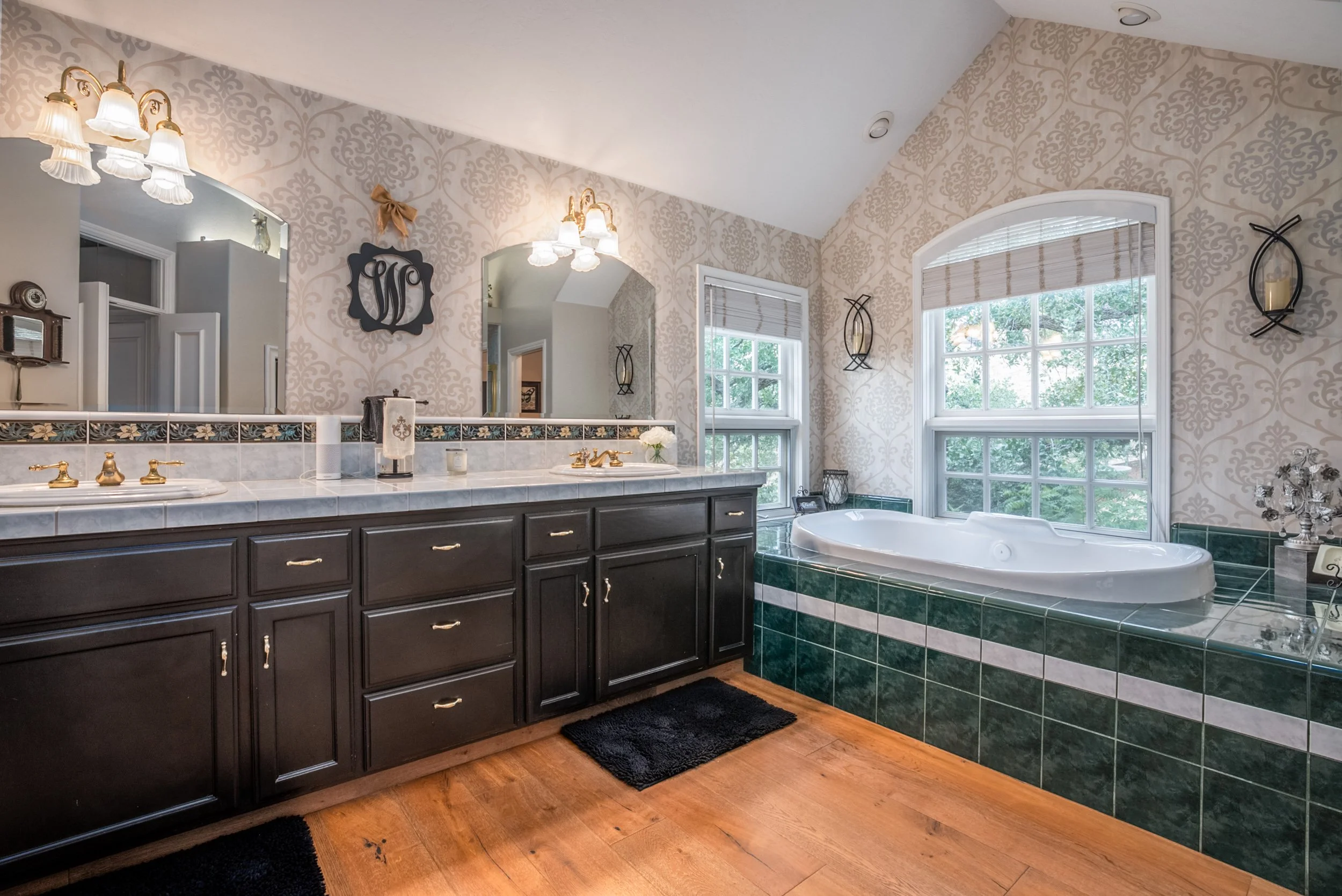 Primary bathroom with dual vanity, brass fixtures, green-tiled soaking tub, and arched window