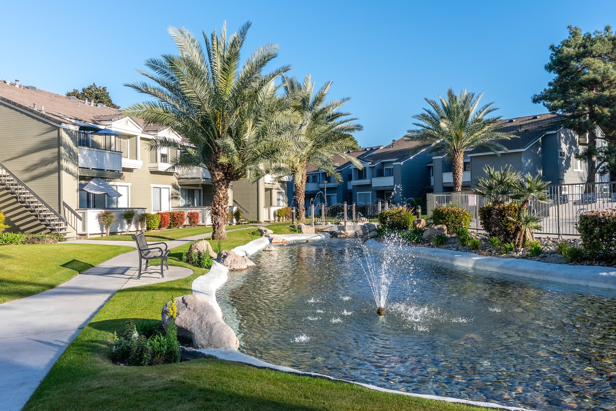 Landscaped apartment community grounds with a fountain pond, palm trees, walking path, and bench