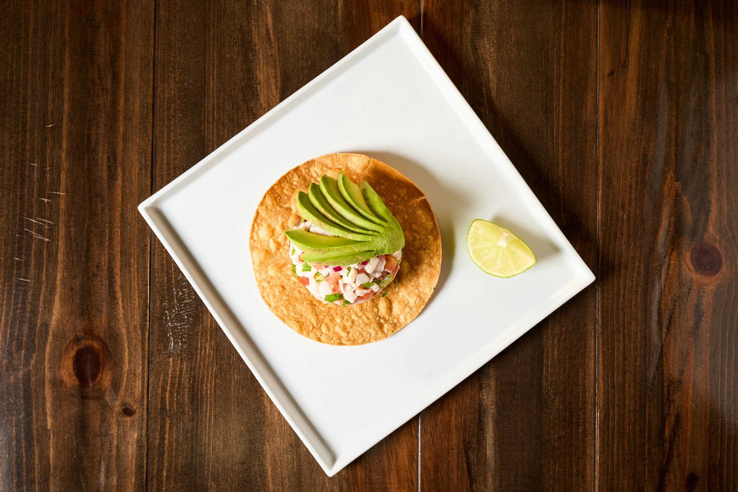 Shrimp ceviche tostada topped with sliced avocado and lime on white square plate — restaurant menu photography by BK Captured"