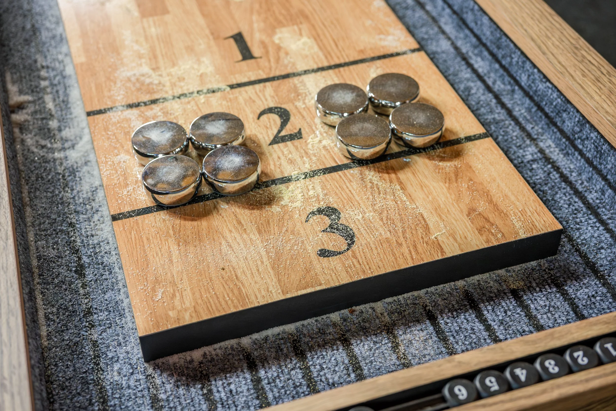 Shuffleboard game with silver pucks on a wooden court, numbers 1 to 3 visible, and powdery residue from play.