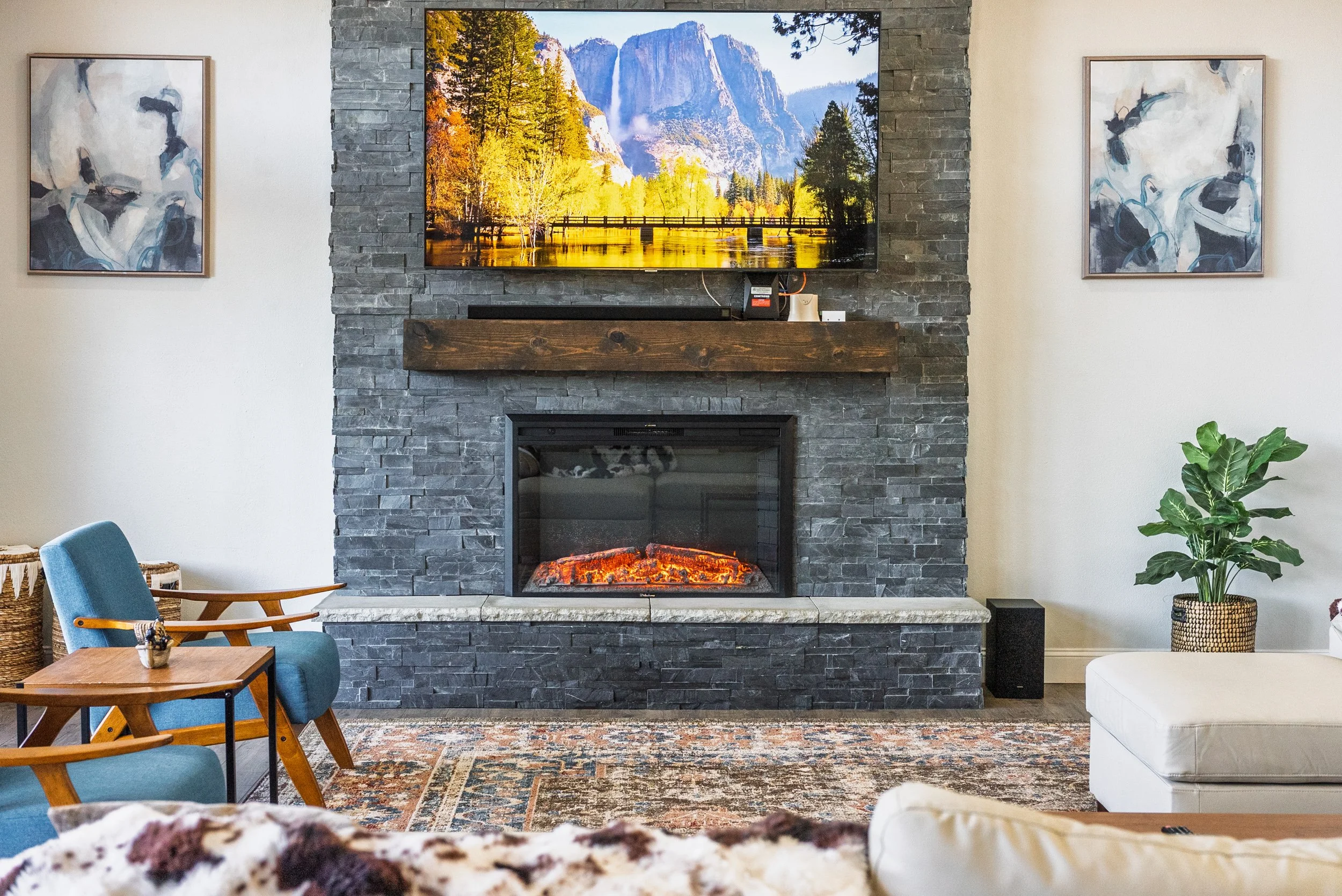 Living room with a stone fireplace and a large wall-mounted TV displaying a mountain and forest scene, flanked by two abstract paintings, with a blue armchair, a small wooden table, and a white sofa. Potted plant on the right.