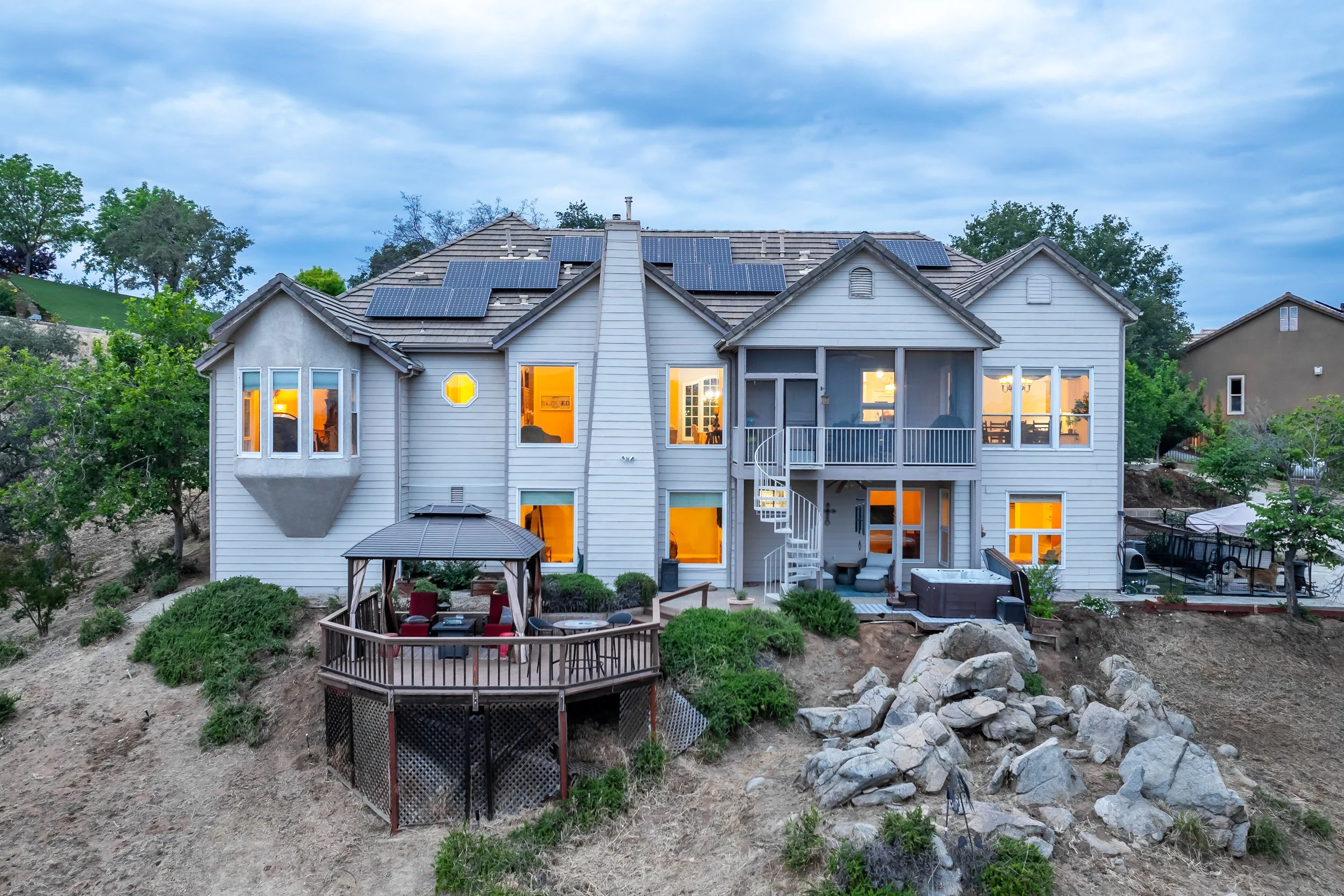 Rear elevation at twilight with solar panels, glowing windows, spiral staircase, and hot tub