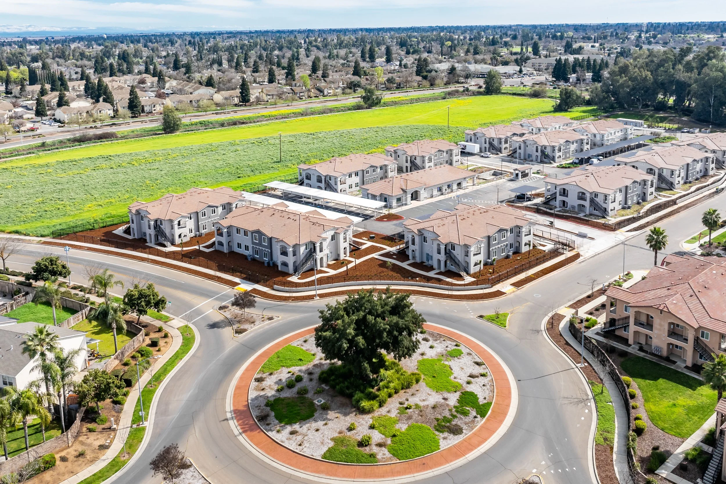 Aerial view of a multifamily apartment community with a landscaped roundabout, surrounded by farmland and neighboring homes