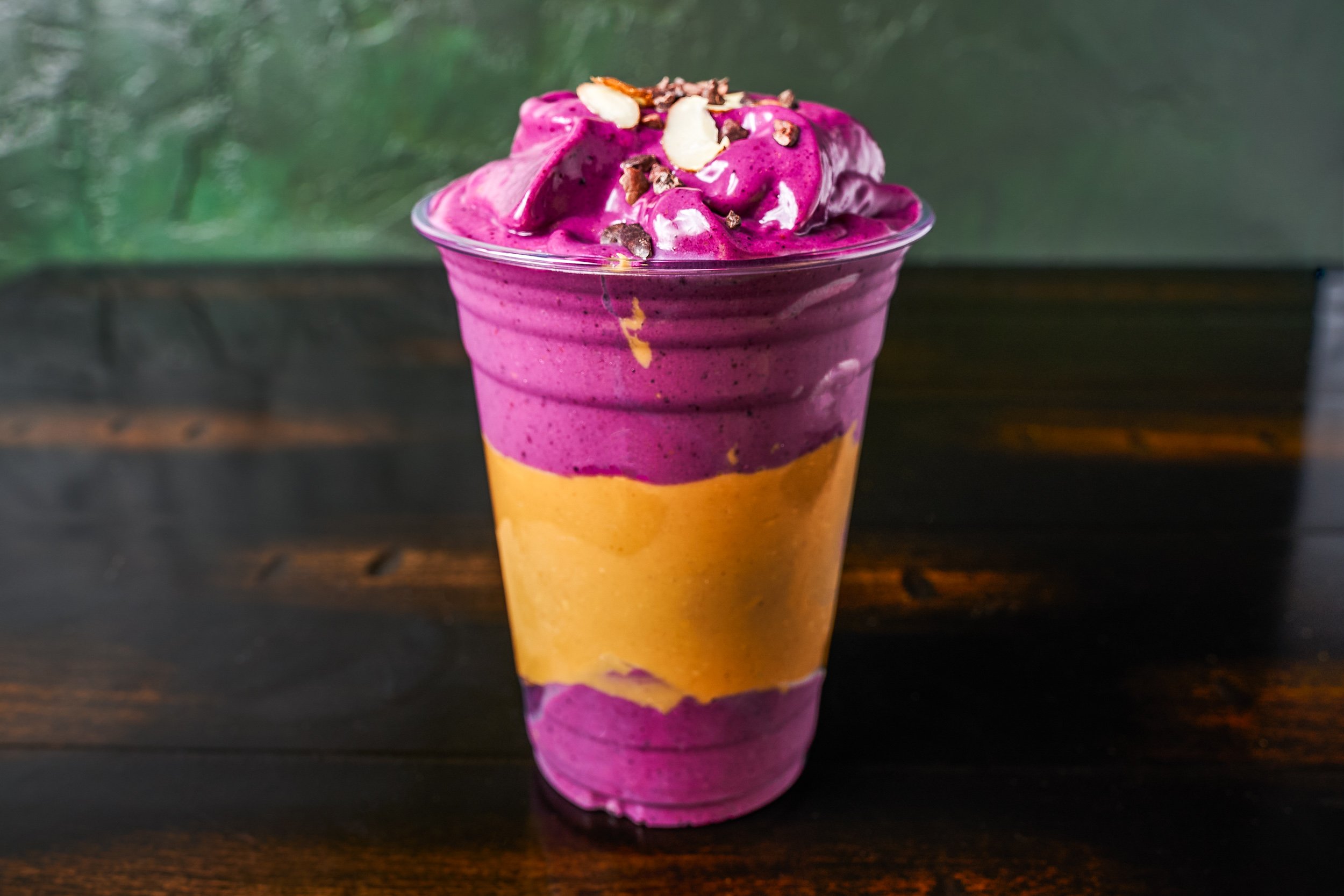 Layered purple and orange peanut butter smoothie topped with almonds and cacao nibs on green background — beverage photography by BK Captured