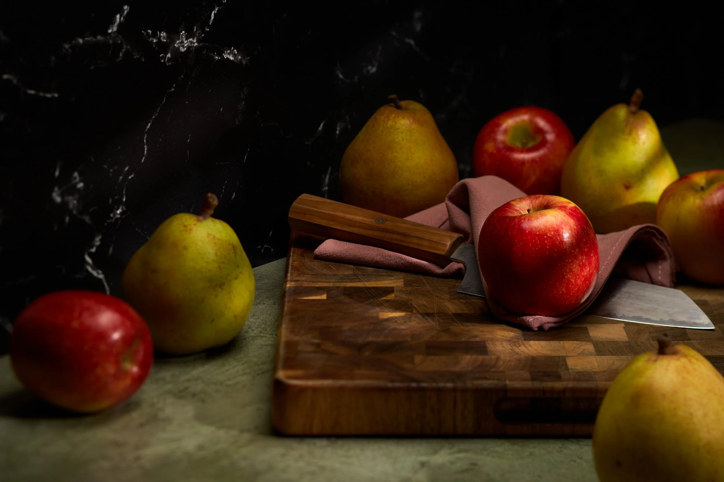 Dark moody styled food photograph of red apples and Bosc pears arranged around a walnut cutting board with chef's knife and linen — fine food photography by BK Captured