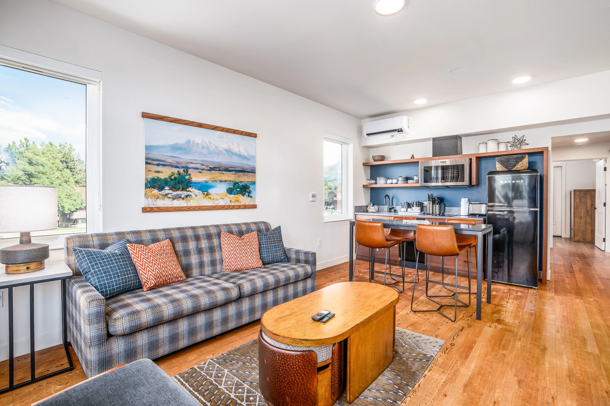 Living room and kitchen area with a plaid sofa, colorful pillows, wooden coffee table, and a view outside the window. The kitchen has blue accents, open shelving, and a black refrigerator.
