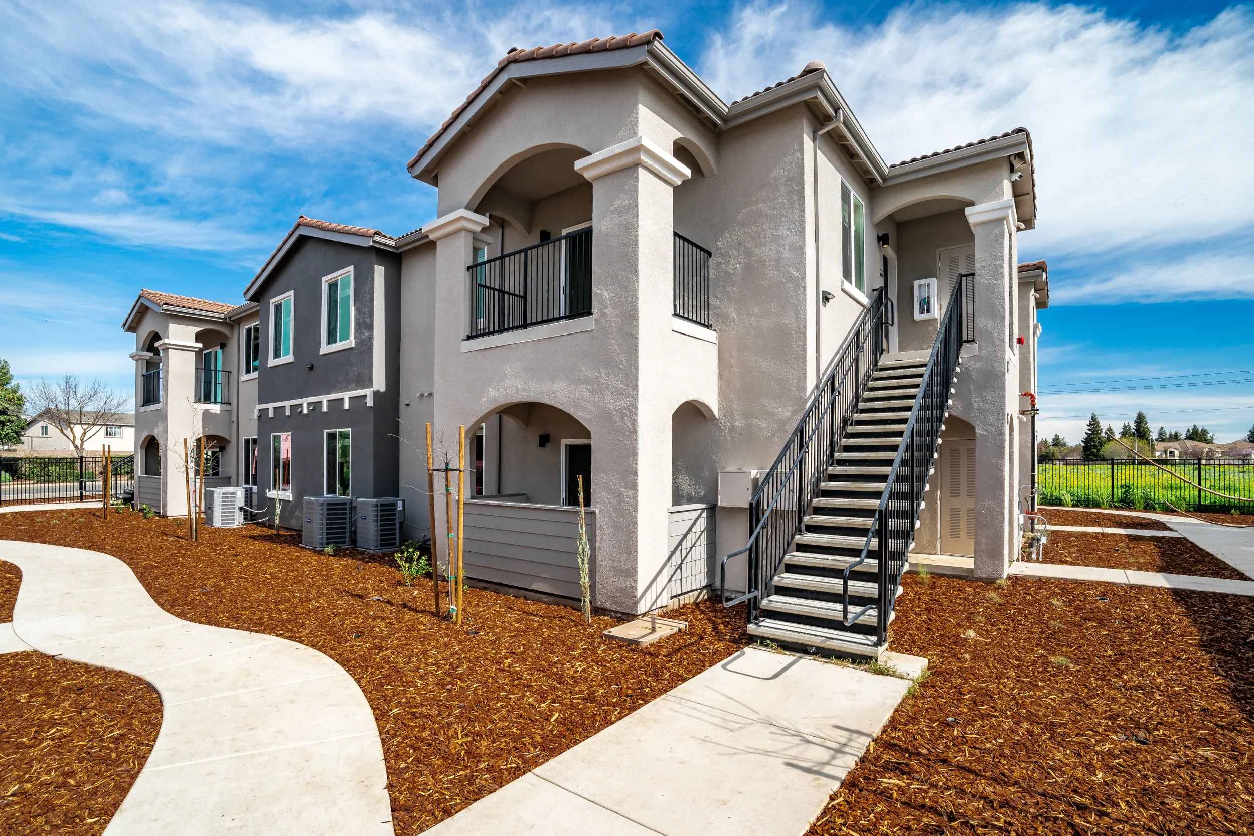 Two-story stucco apartment building with tile roof, exterior staircase, and fresh landscaping under a blue sky