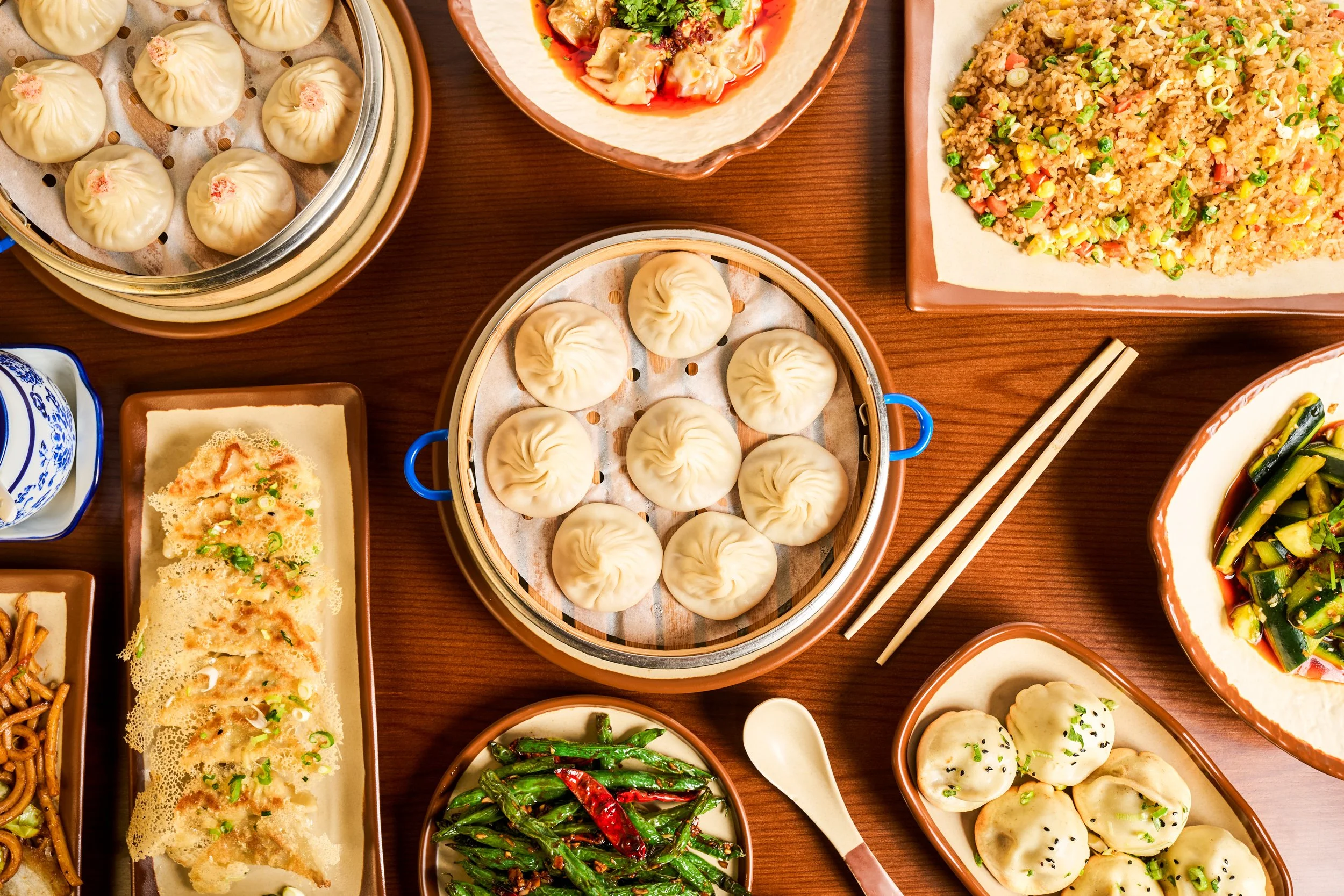 Overhead flatlay of Chinese restaurant spread with soup dumplings, scallion pancake, fried rice, green beans, and steamed buns on warm wood surface — restaurant photography by BK Captured