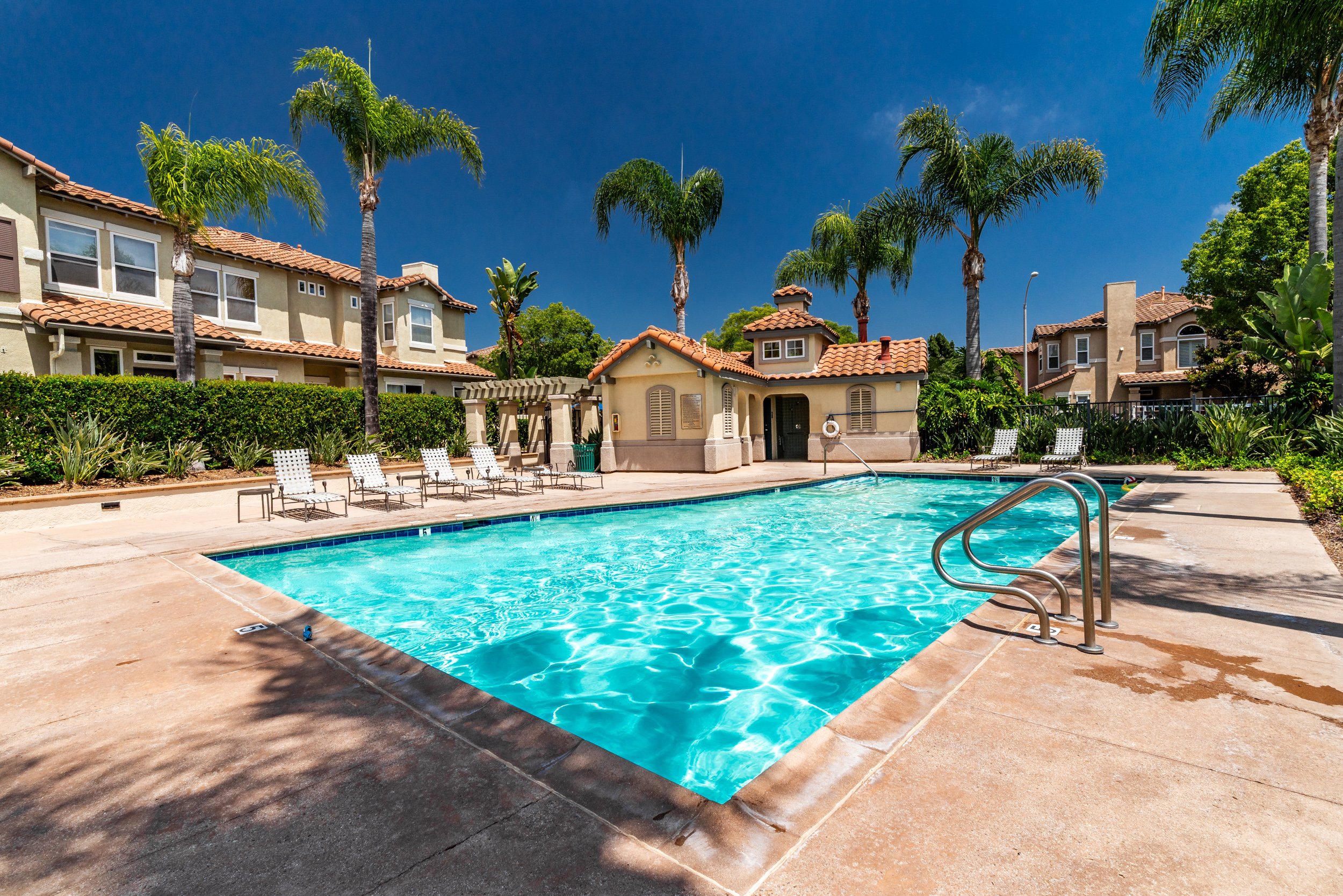 An outdoor swimming pool surrounded by lounge chairs, lush greenery, and multi-story residential buildings with red-tile roofs under a clear blue sky.