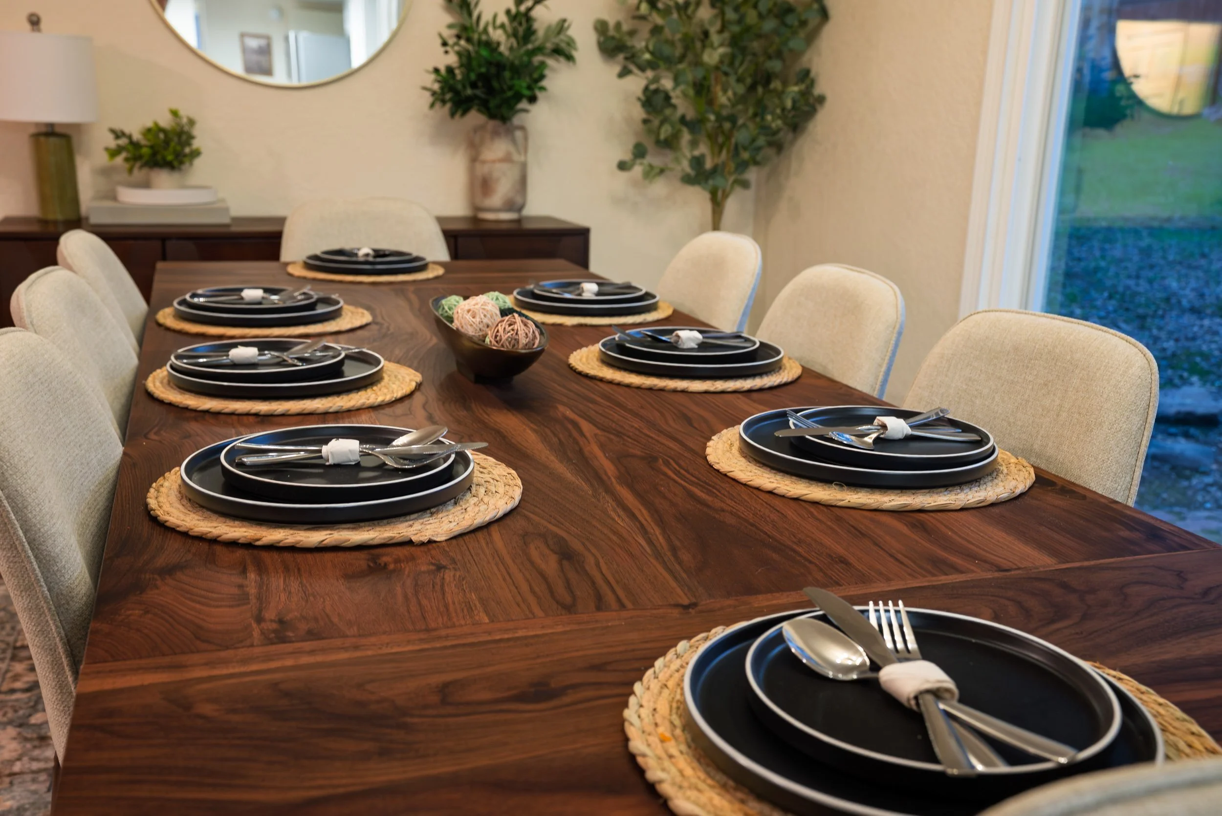 A dining room table set with black plates, silverware, and beige napkins on a woven placemat, with a bowl of decorative yarn balls in the center. The table is surrounded by cream-colored chairs, and there's a mirror and green plants on the wall in th