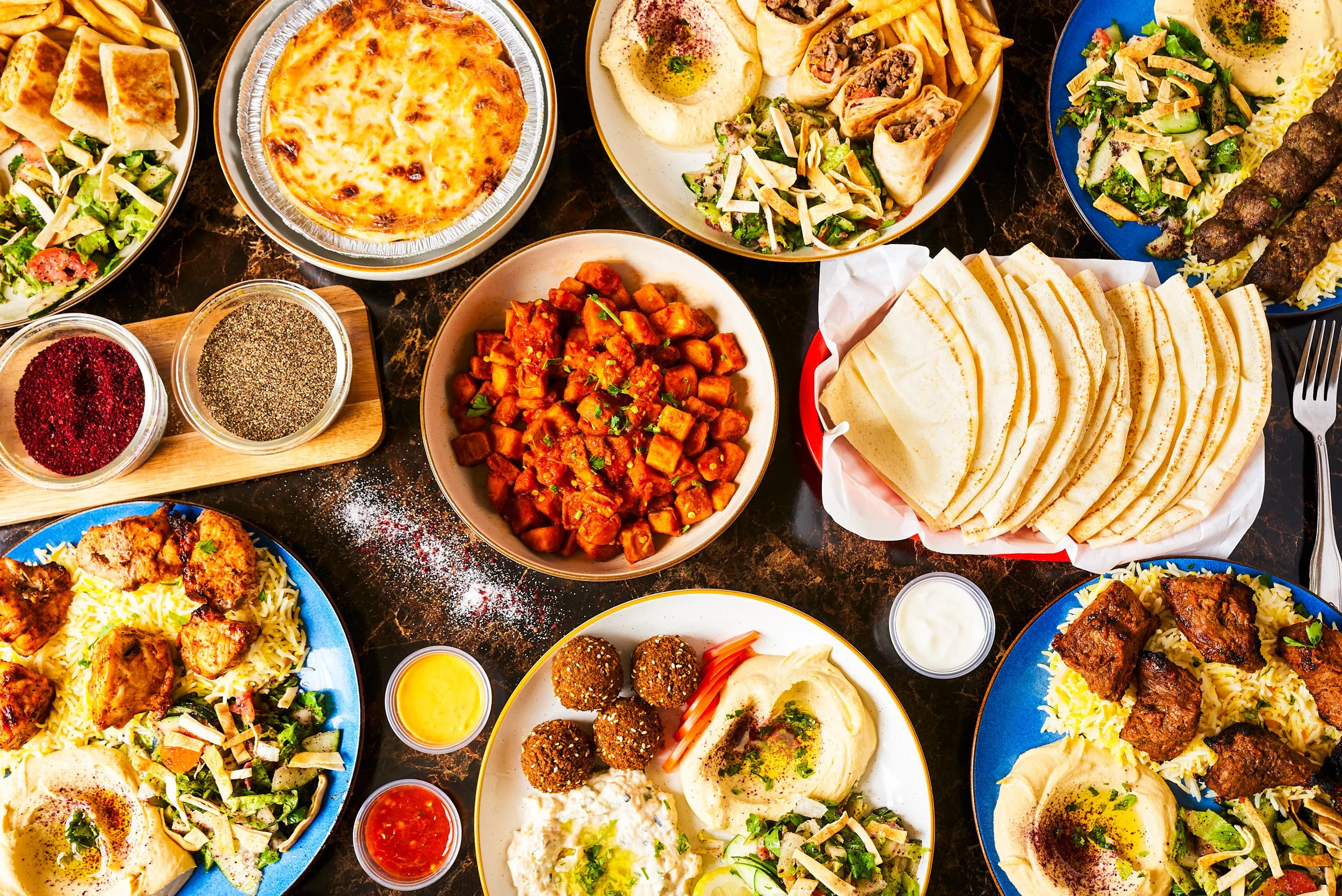 Overhead flatlay of Middle Eastern restaurant spread with hummus, falafel, pita bread, kabobs, and spiced potatoes on dark marble — restaurant photography by BK Captured