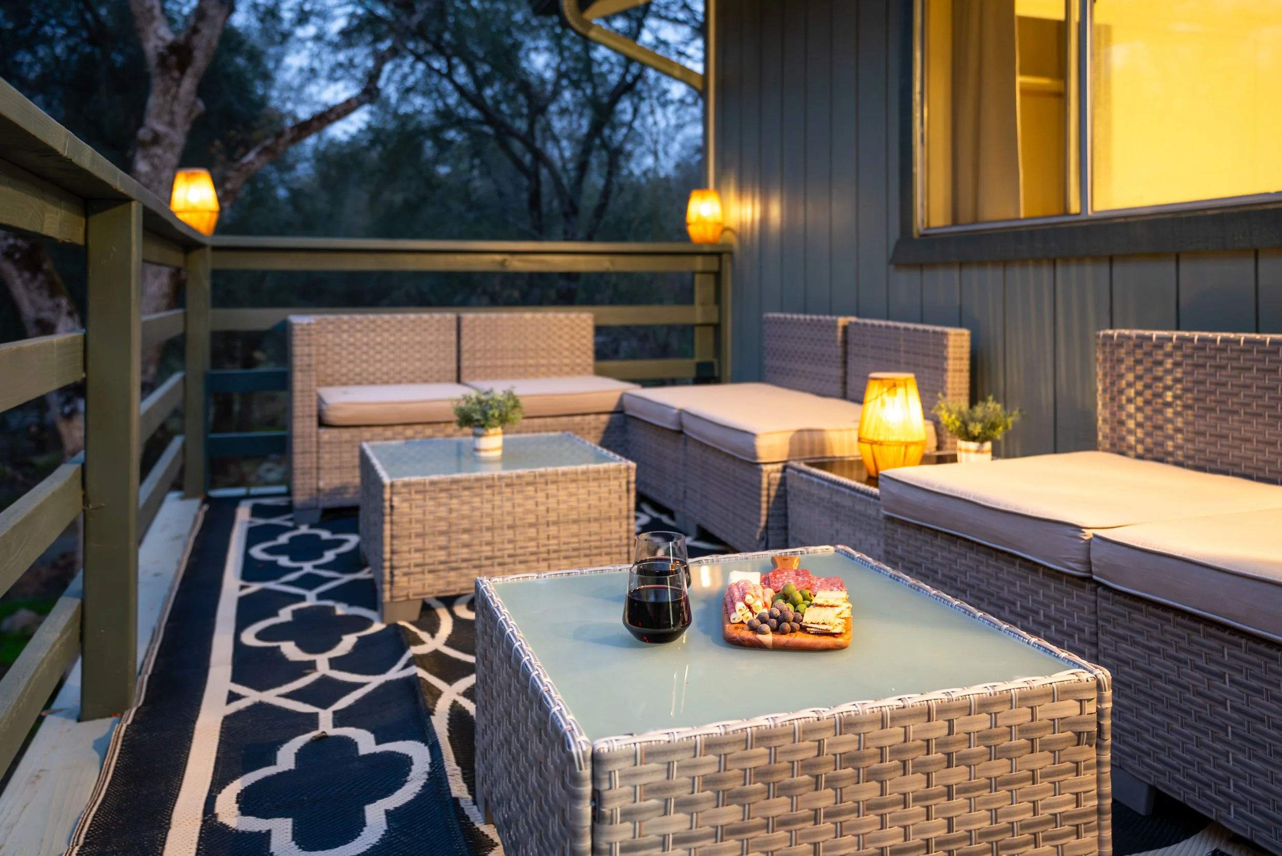 Outdoor balcony with wicker furniture, night time setting, lit lanterns, a table with wine glasses and snacks, and trees in the background.