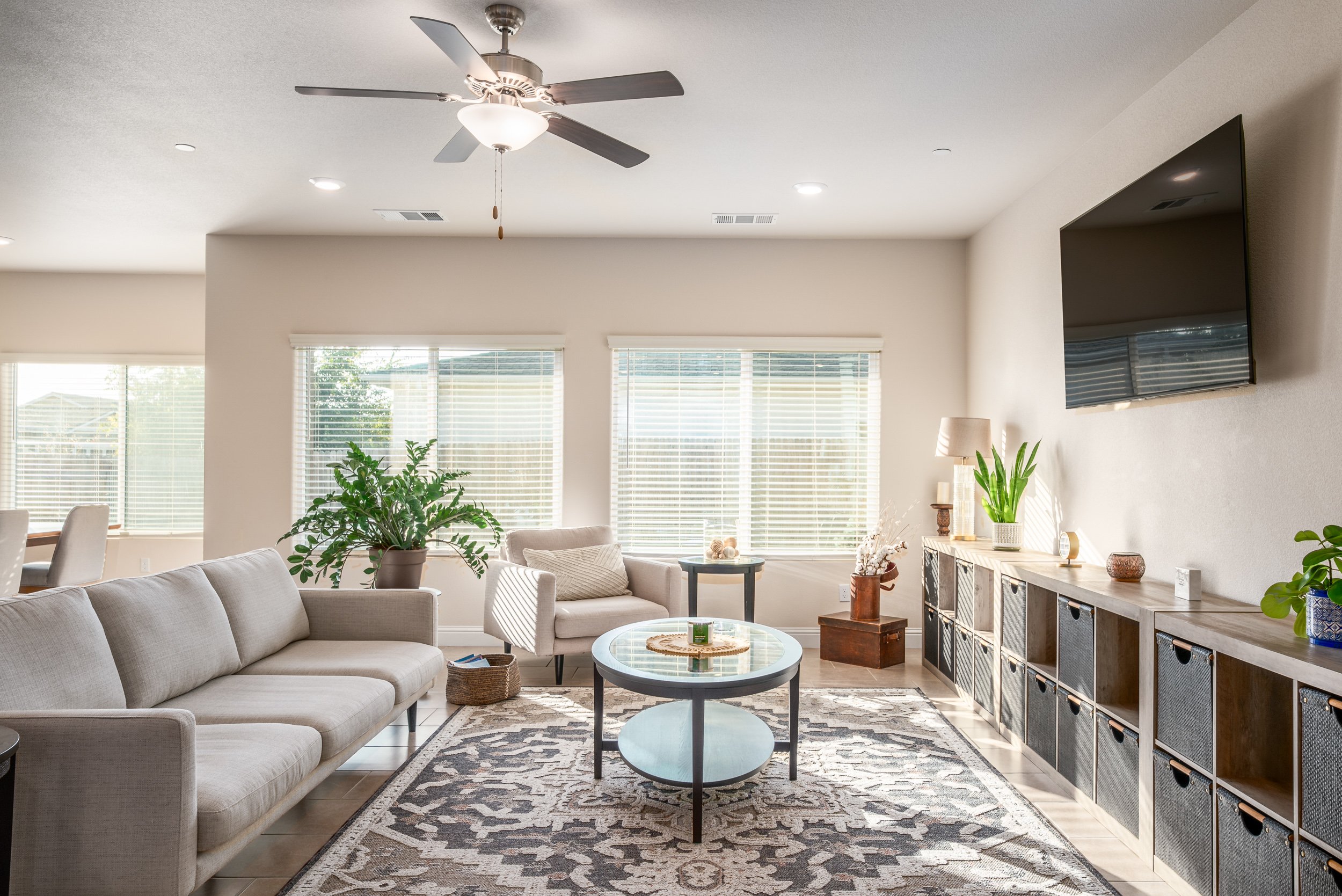 Bright staged living room with a neutral sofa, ceiling fan, cube storage, and large windows