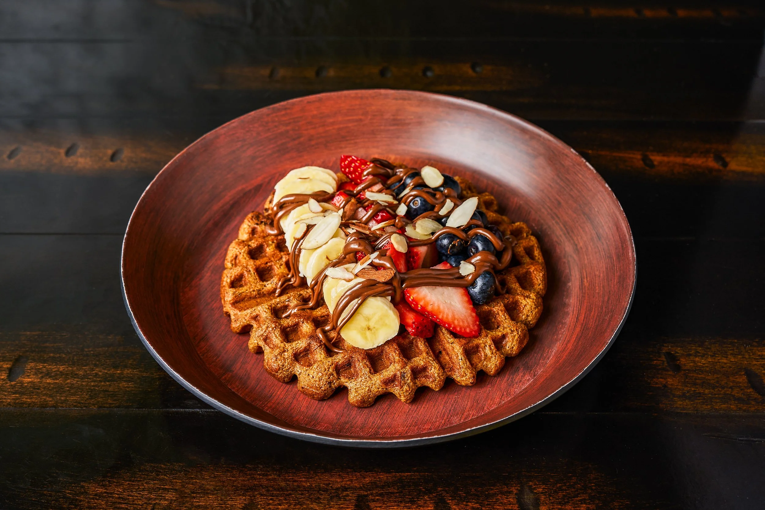 Whole grain waffle topped with Nutella drizzle, fresh strawberries, blueberries, banana, and sliced almonds on dark wood — restaurant food photography by BK Captured