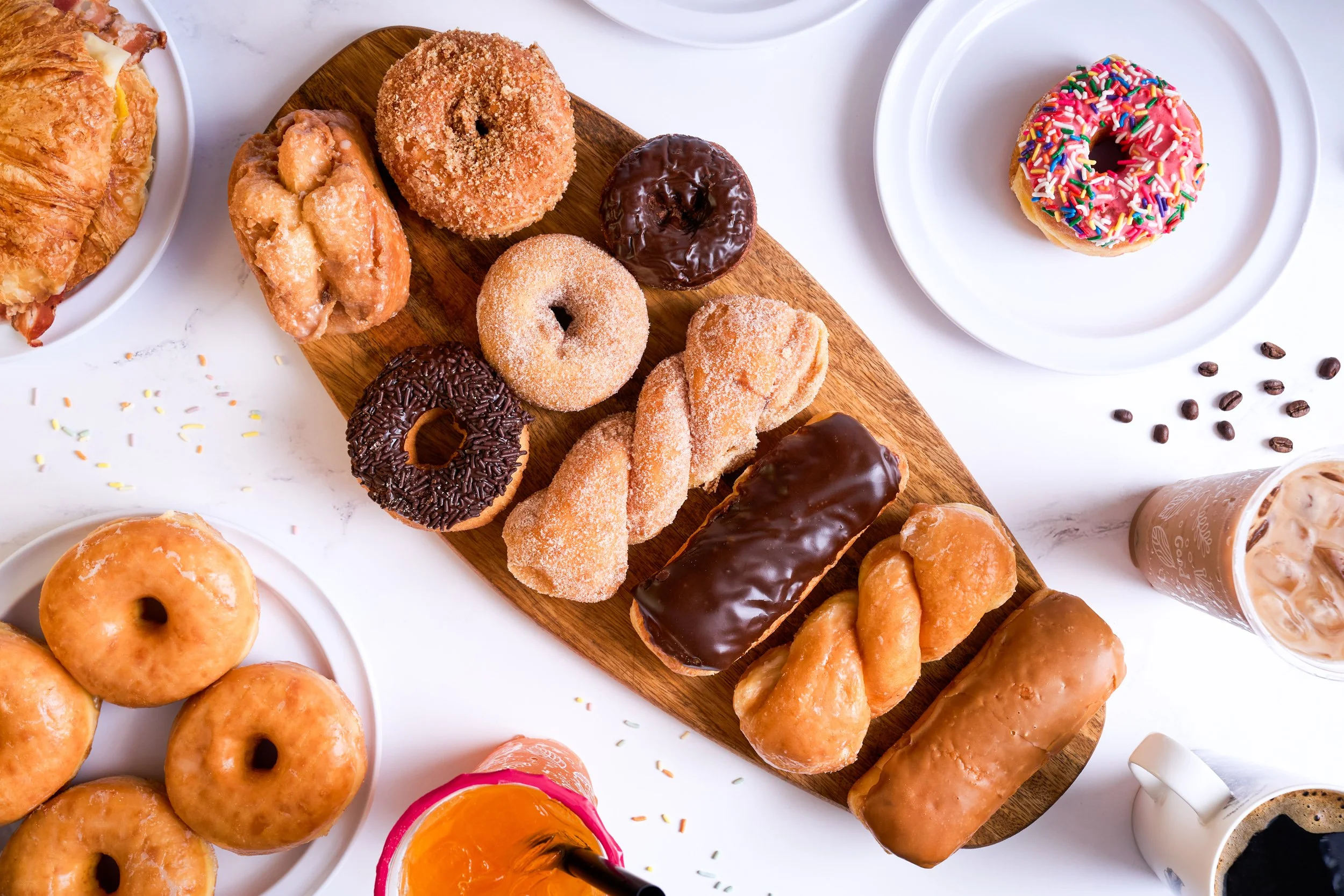 Overhead flatlay of assorted donuts on wooden board with iced coffee and coffee beans on white marble — bakery food photography by BK Captured