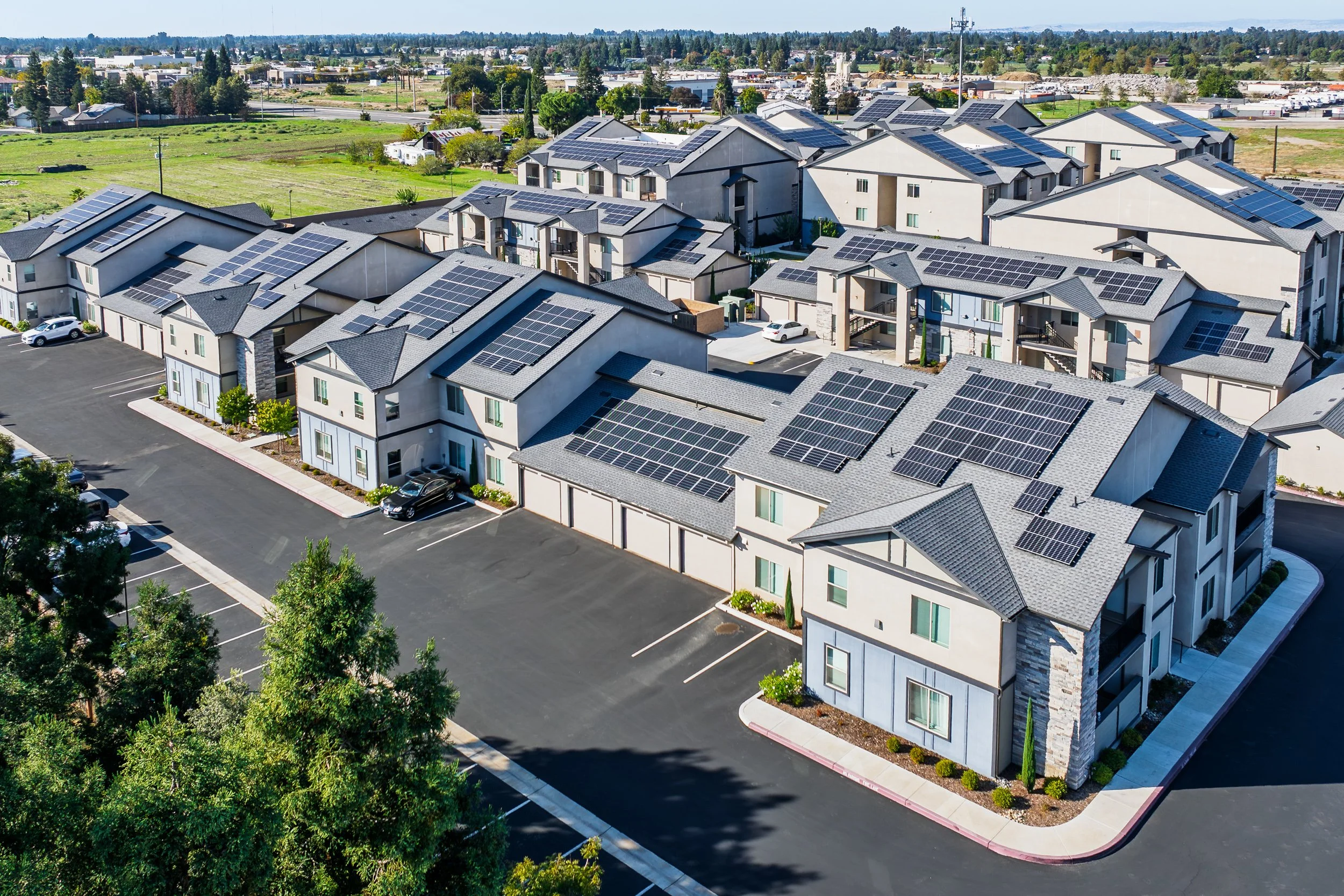 Aerial view of a solar-paneled multifamily apartment complex with parking and open fields beyond