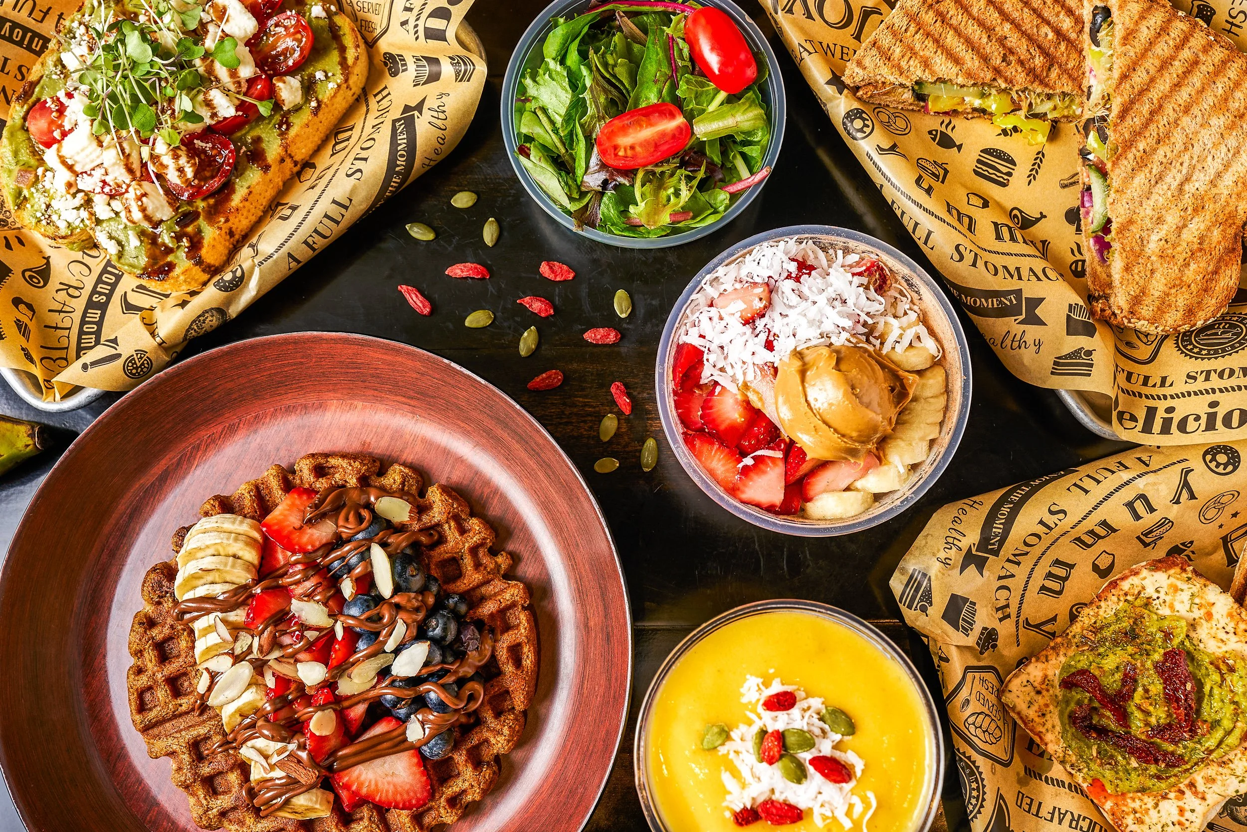 Overhead flatlay of Go Greek Fresno restaurant menu including Nutella waffle, avocado toast, acai bowl, panini, and mango smoothie on dark surface — restaurant photography by BK Captured