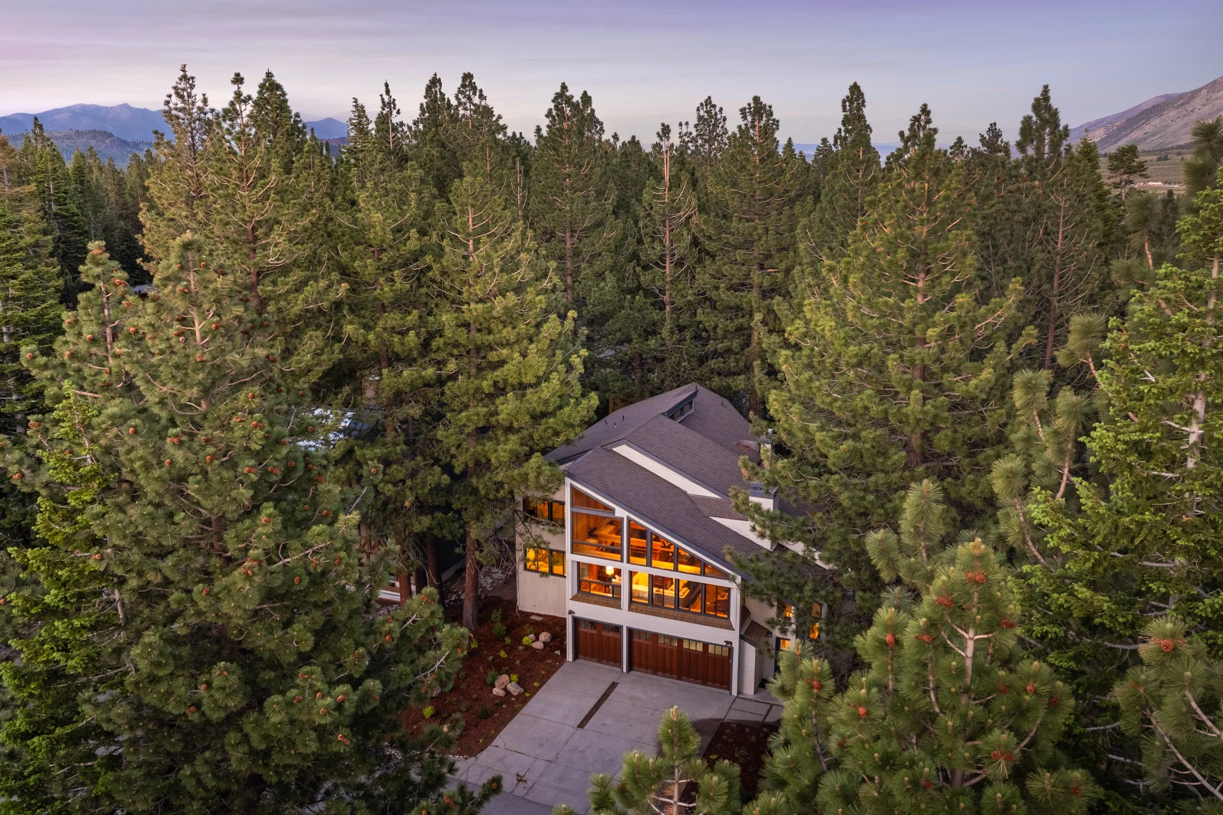 Aerial of a glass-fronted mountain home glowing at twilight, nestled in tall pines with distant peaks